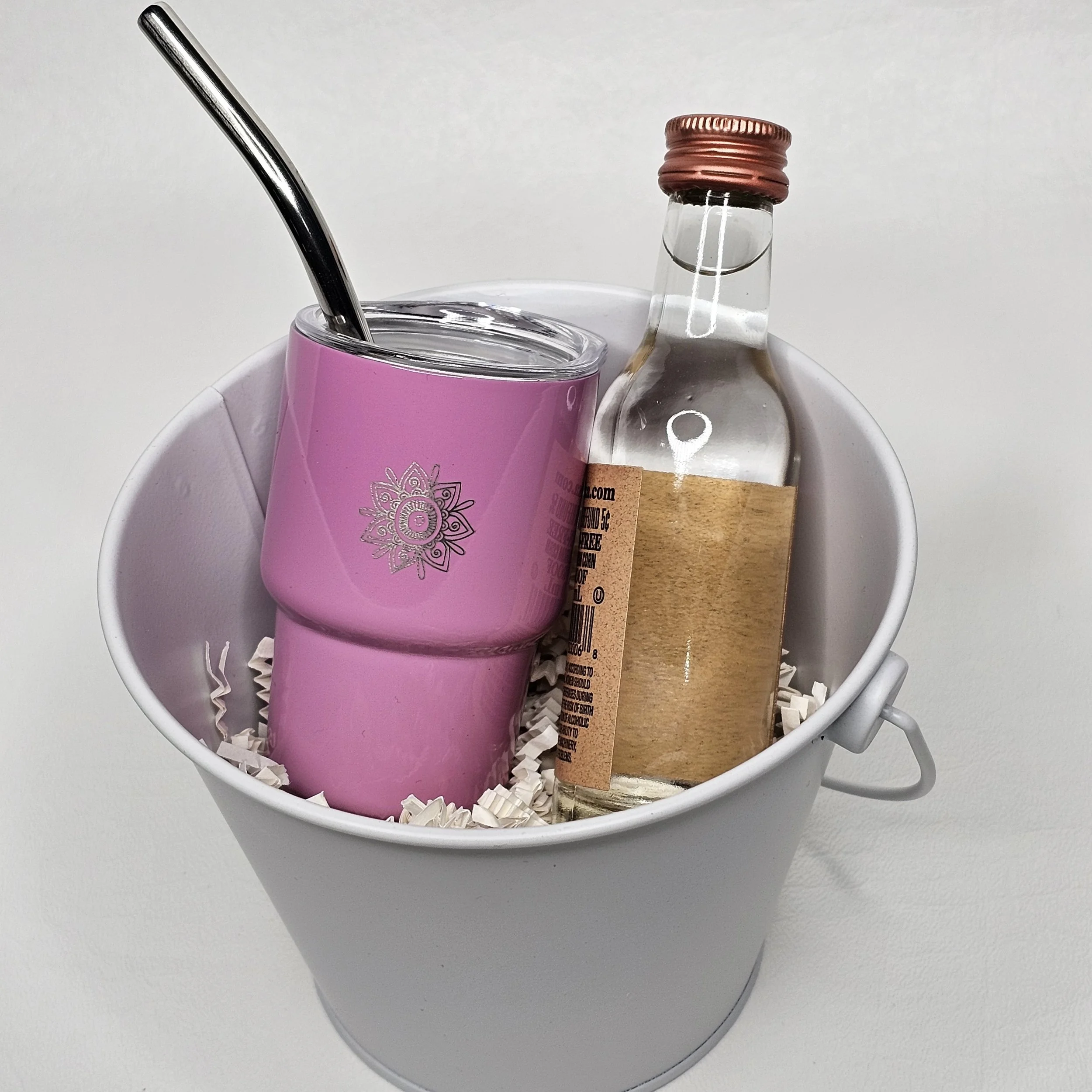 A white bucket holding a pink tumbler with a straw and a small glass bottle with a brown label and pink cap, resting on shredded paper.