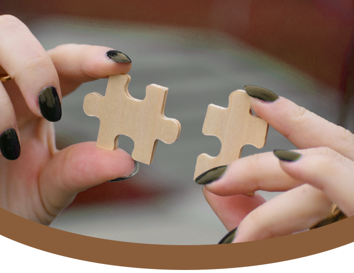 Two hands with black painted nails holding two separate wooden puzzle pieces, about to fit them together.