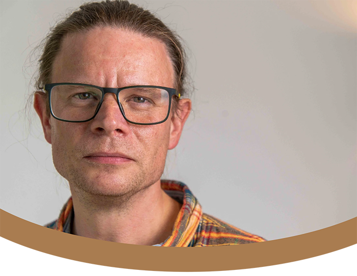Gavin Demurger-Jones, Counsellor, Psychotherapist and Practice Director at held. Wearing a plaid shirt, looking serious, standing against a plain light-colored wall.