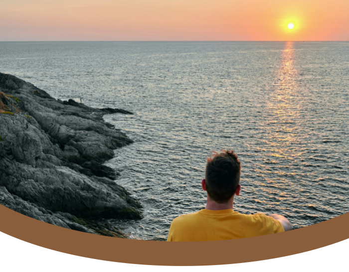 A person with short dark hair wearing a yellow shirt sitting on rocky coastline watching sunset over the ocean.