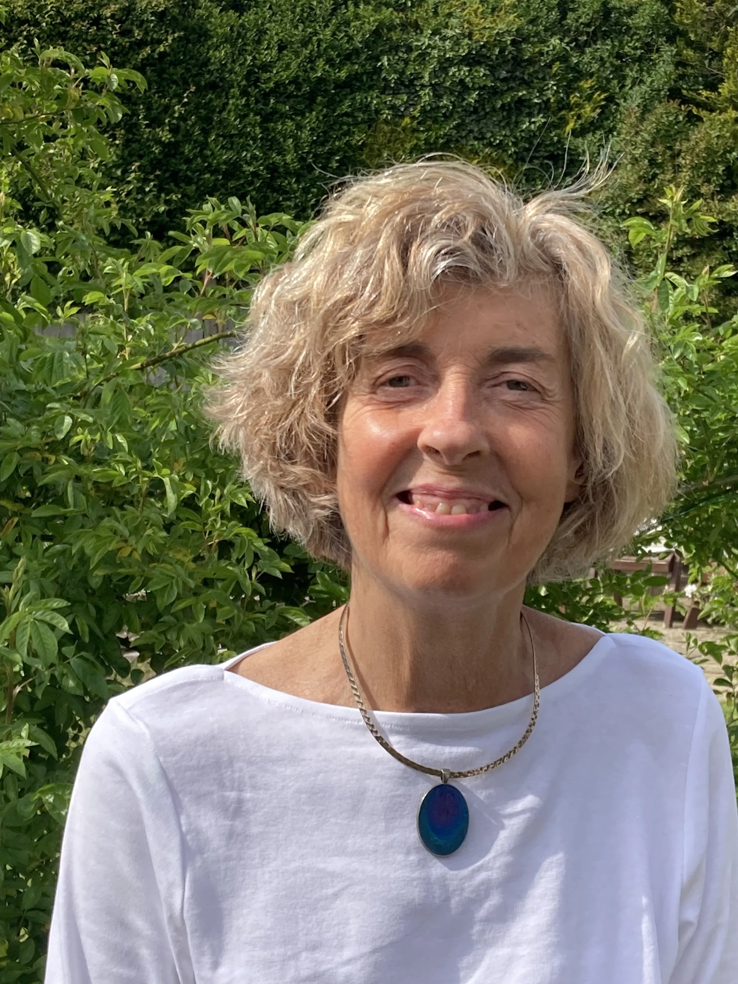 Ali Cardosi, Counsellor and Psychotherapist at held, with curly blonde hair, wearing a white top and a necklace with a large blue pendant, standing outdoors with green leafy bushes in the background.