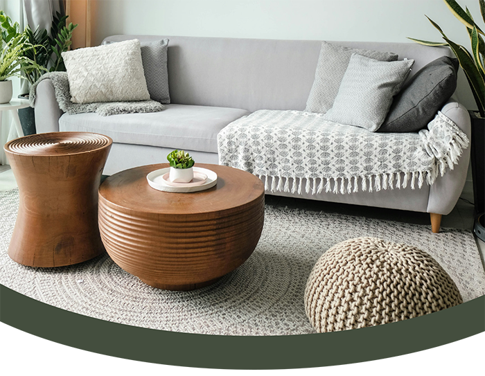 Living room with a light gray sofa decorated with gray and white pillows, a knitted blanket, and a throw. In front of the sofa are two wooden coffee tables and a beige knitted pouf. The room has a round woven rug and green houseplants on the side.