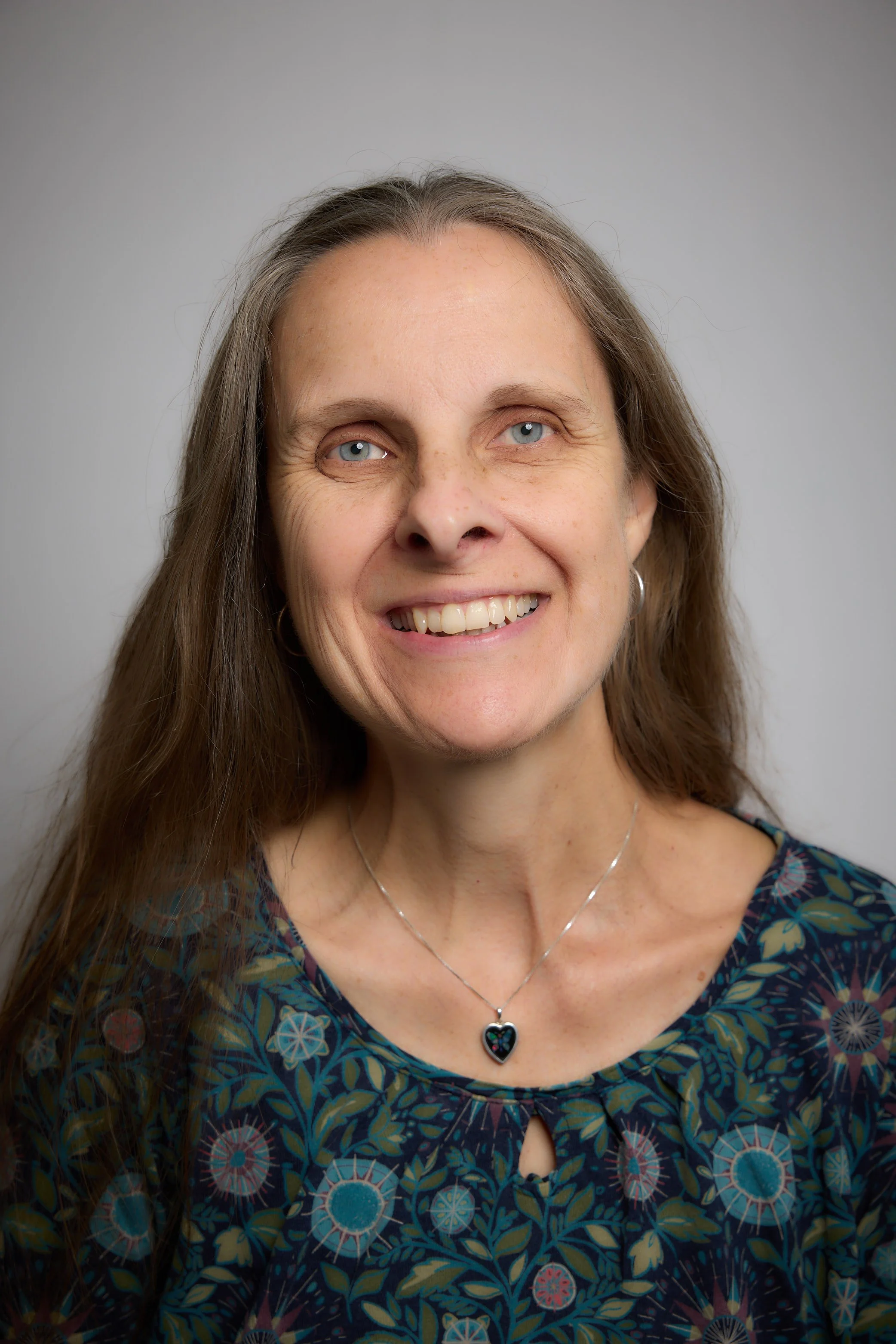 Karen Hanson, Counsellor and Psychotherapist at held, with long brown hair, blue eyes, and a necklace with a heart-shaped pendant, wearing a blue floral patterned top.