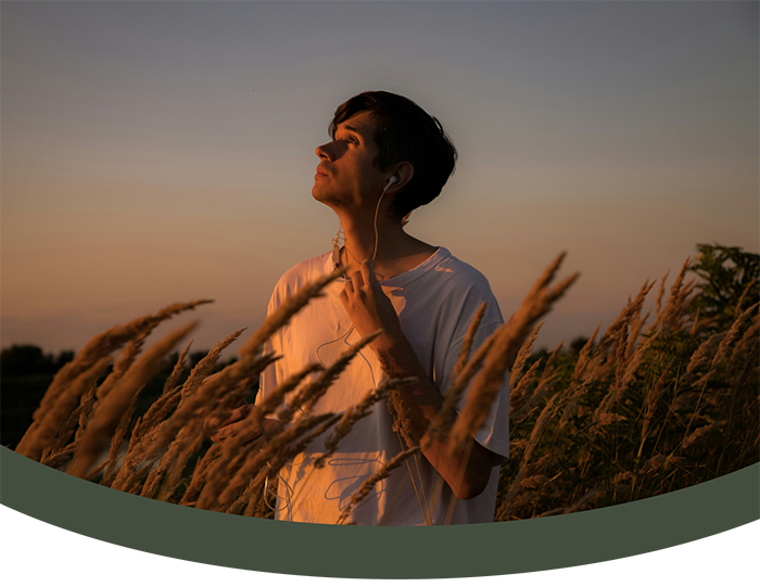 Young man listening to music with earphones in a field of tall grass during sunset.