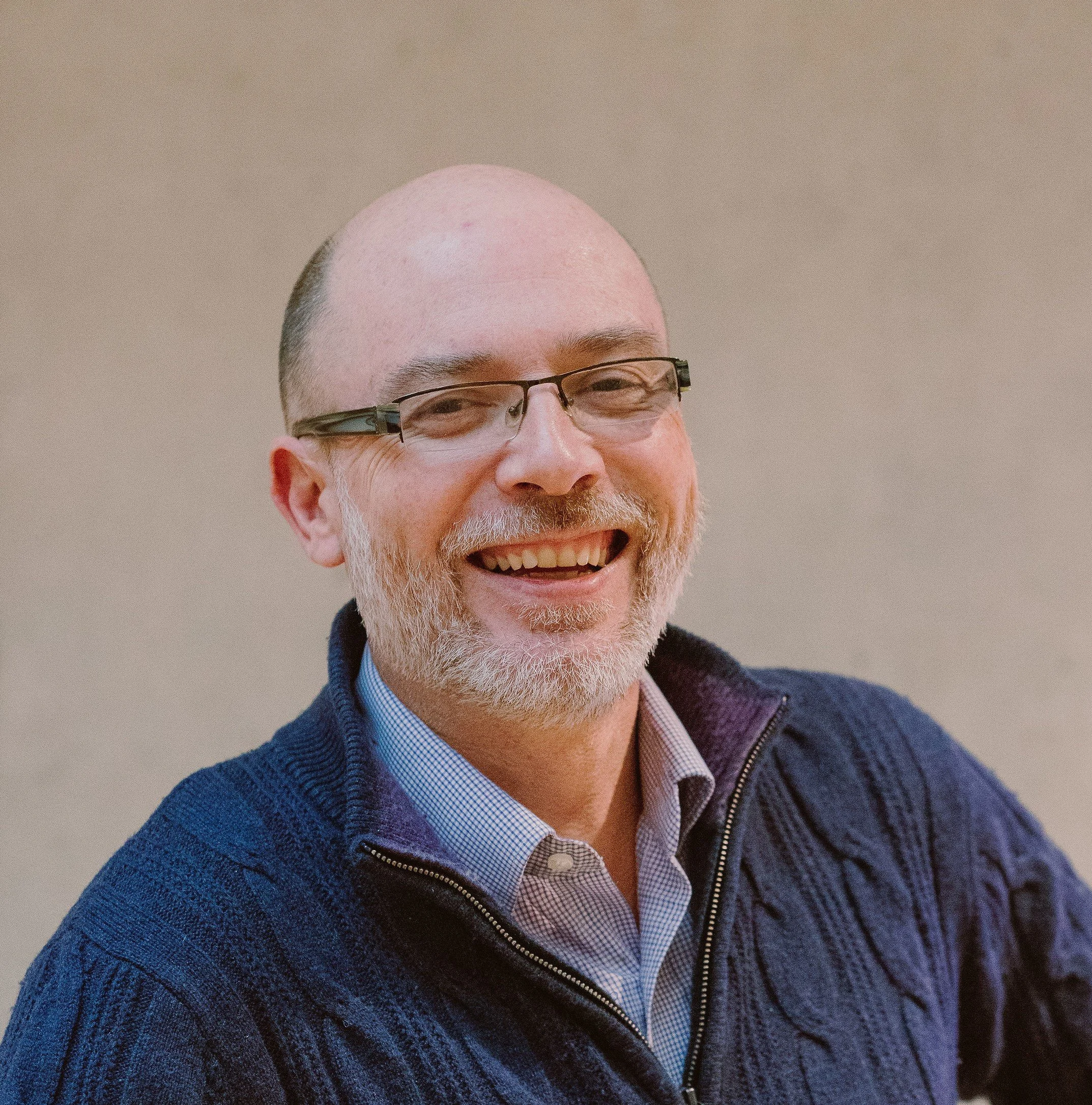 Jeremy Cripps, Clinic Psychologist at held, with glasses, white beard, wearing a blue knit sweater and a collared shirt, background blurred.