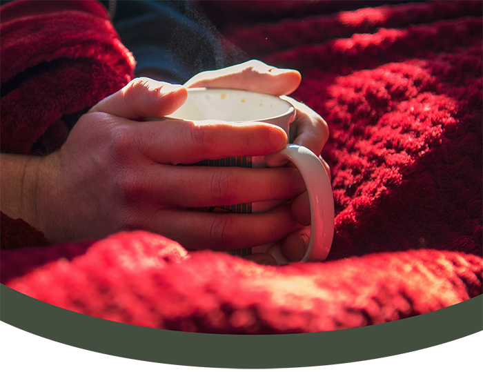 Person holding a mug with both hands, sitting on a red textured blanket or rug.