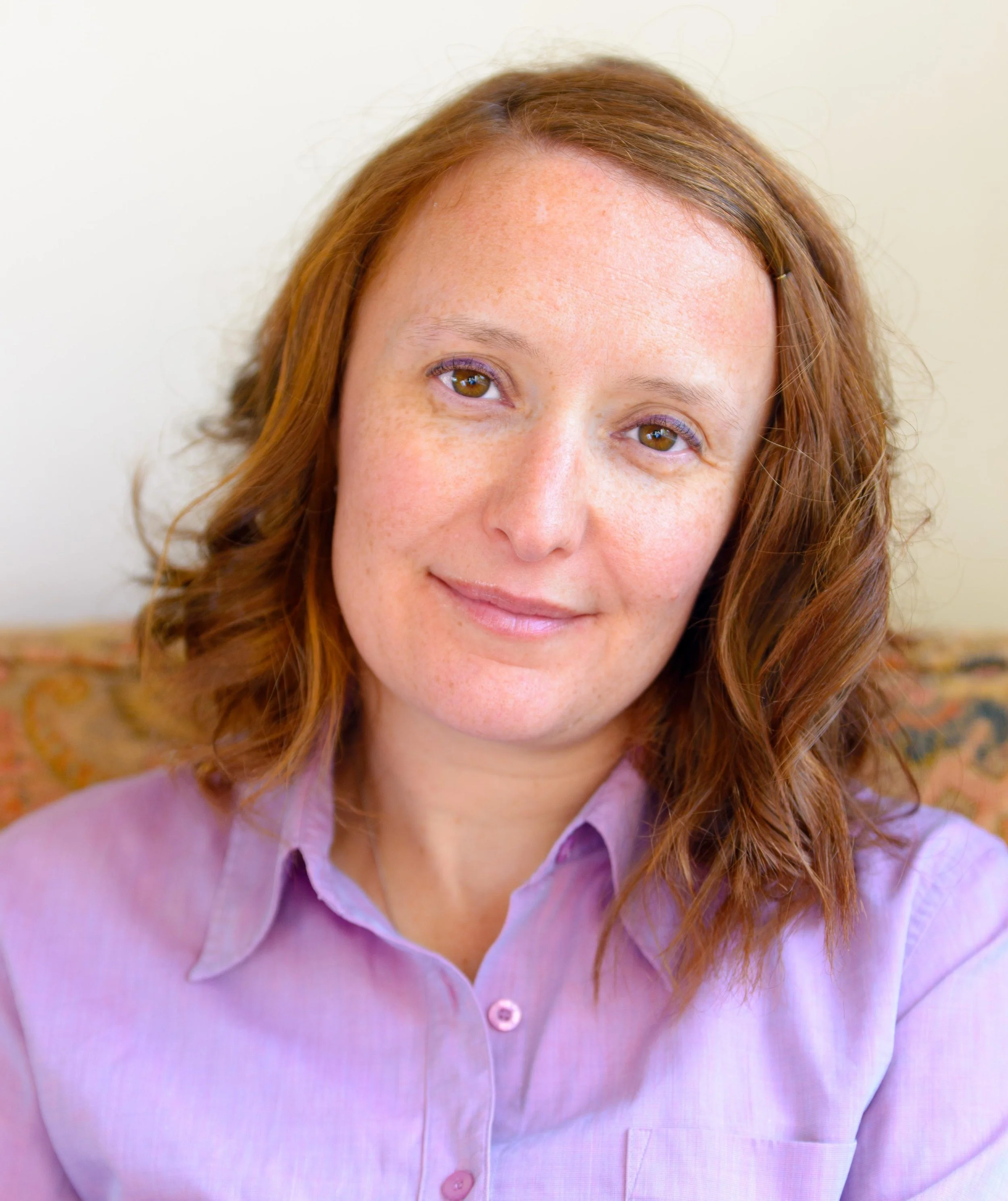 Elina Demurger-Jones, Counsellor and Psychotherapist at held, with red hair, freckles, wearing a light purple collared shirt, smiling softly against a neutral background.