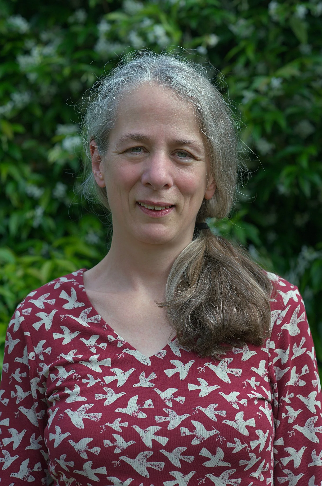 Abigail Warren, Counsellor and Psychotherapist at held, with gray hair, wearing a red top with white bird print, standing outdoors in front of green foliage.