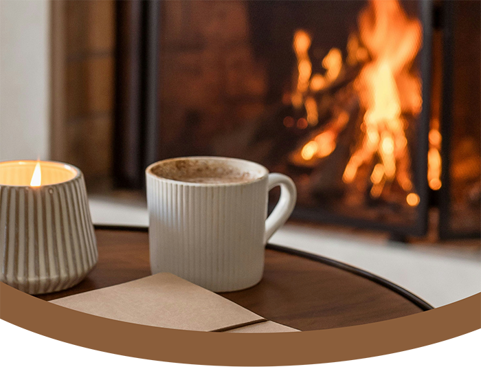 A cozy scene with a lit candle, a mug of hot chocolate or coffee, and a notebook on a wooden table in front of a lit fireplace.