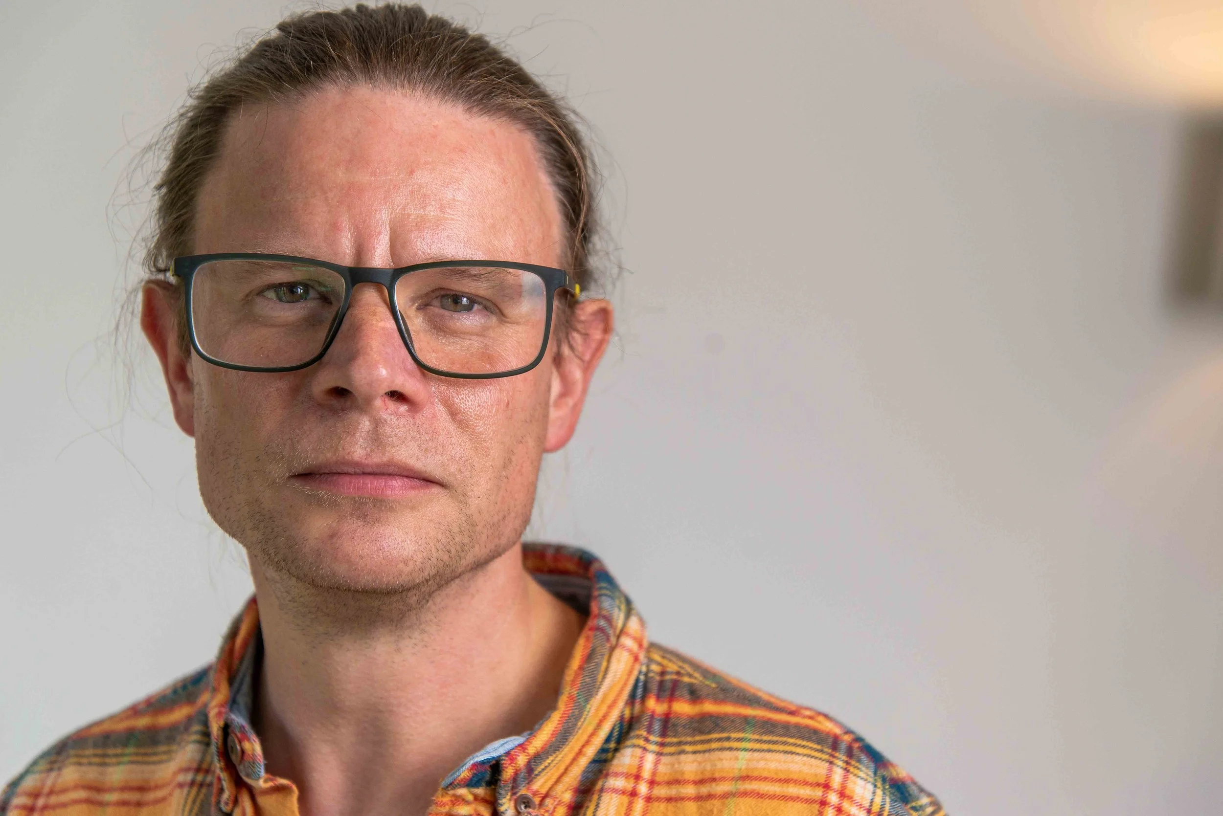 Gavin Demurger-Jones, Counsellor, Psychotherapist and Practice Director at held. Wearing a plaid shirt, looking serious, standing against a plain light-colored wall.