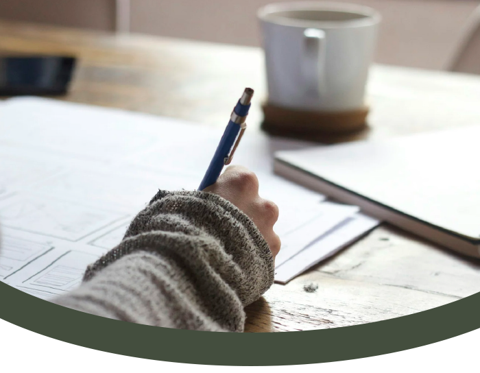 A hand in a striped sweater holding a pen over sheets of paper on a wooden table, with a coffee mug and a closed notebook in the background.