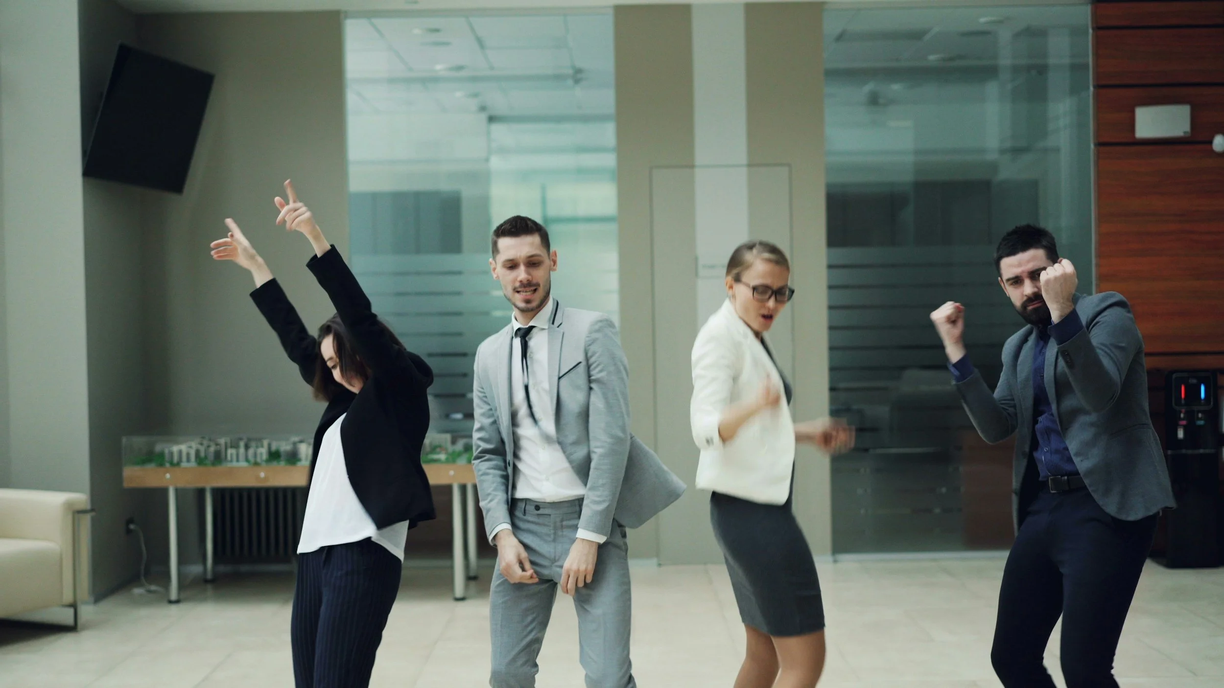 Four young adults in business attire dancing and celebrating in an office lobby.