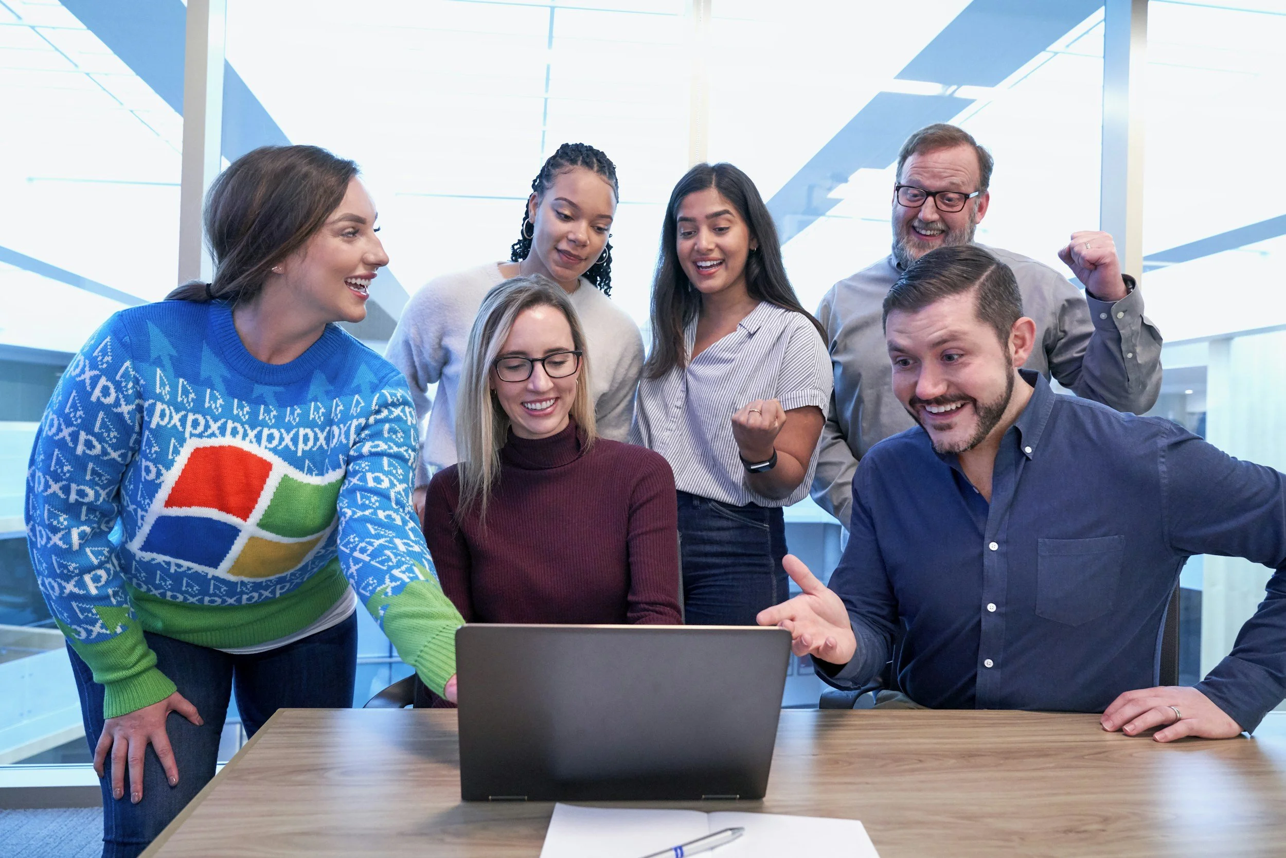 Group of six people gathered around a laptop, smiling and celebrating a successful moment in a modern office setting.