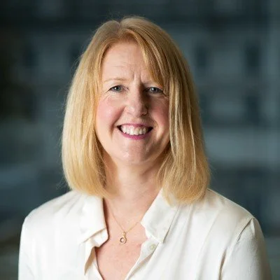 A picture of me with shoulder-length blond hair smiling, wearing a white blouse, in front of a blurred cityscape background.
