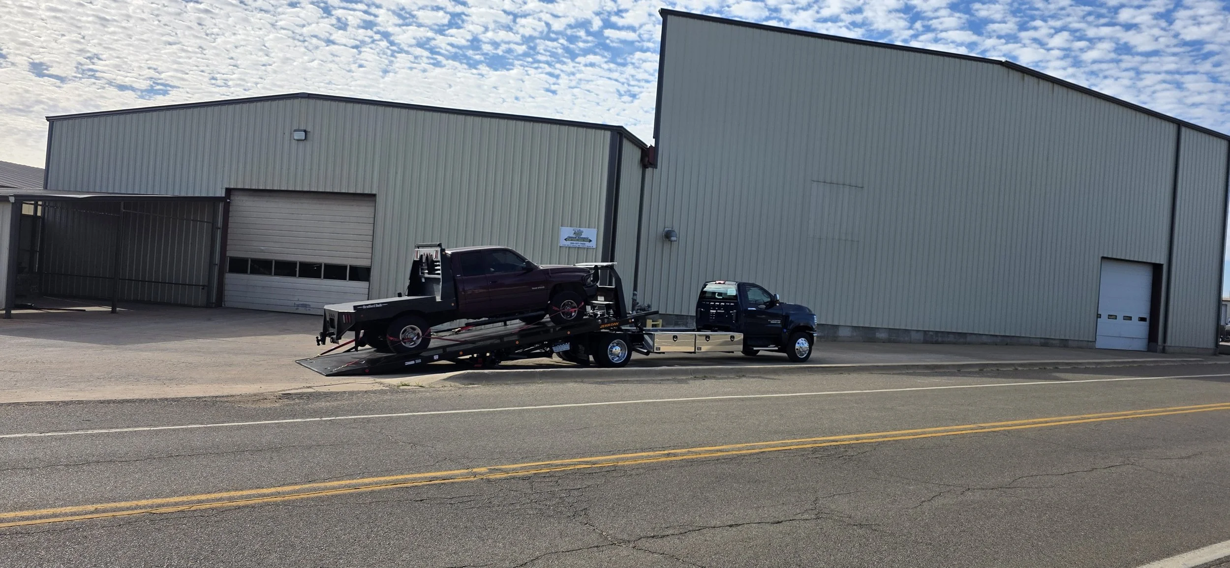 This image shows a flatbed tow truck parked along the side of a paved road in front of a large industrial metal building. The truck is in the process of loading a dark-colored pickup onto its tilted rollback bed, with the rear of the bed lowered and 
