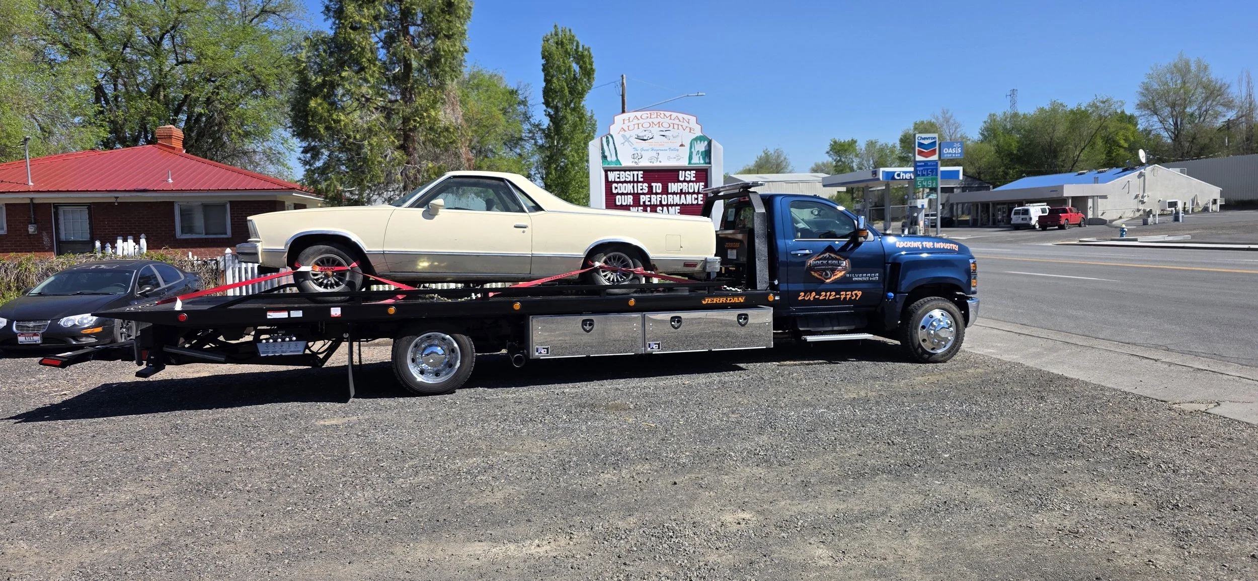 This image shows a flatbed tow truck parked on a gravel lot, carrying a classic cream-colored chevy El Camino. The truck appears to be a heavy-duty rollback with a long black bed, secured with bright red tie-down straps holding the vintage car firmly