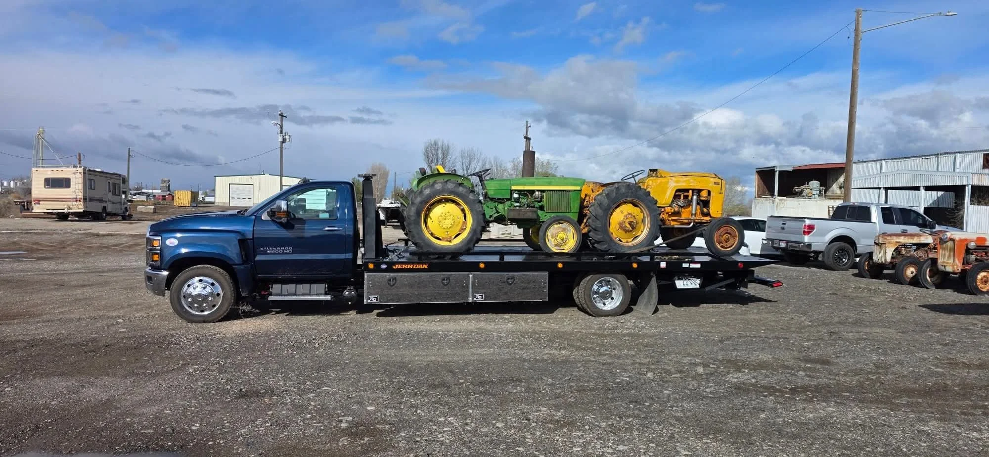 This image shows a heavy-duty flatbed tow truck parked on a gravel lot, hauling two vintage tractors. The truck, a dark blue rollback, has a black bed with metal toolboxes along the side and appears built for medium to heavy transport jobs.
On the be