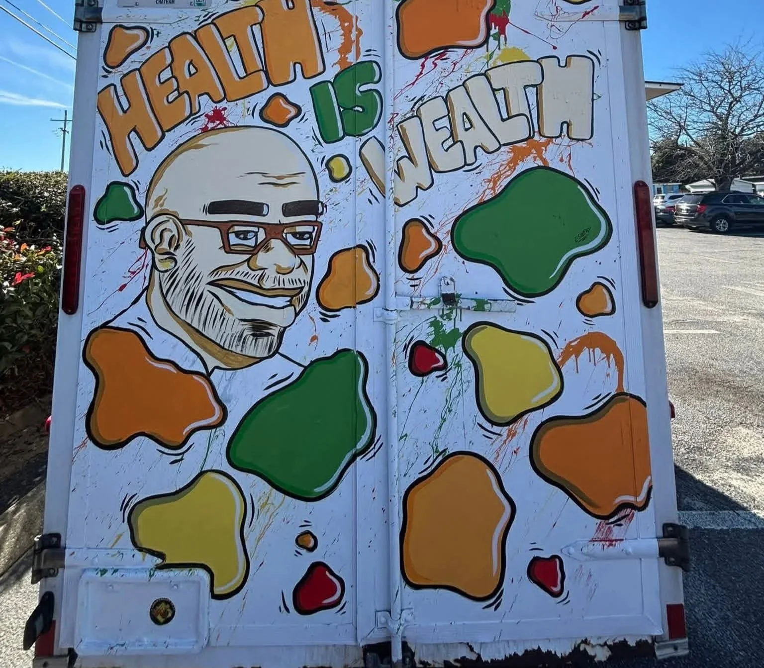 Colorful street art on a vehicle depicting a smiling man with glasses and a beard, surrounded by abstract shapes in green, yellow, orange, and red, with the words 'Healthy is Wealth' painted at the top.