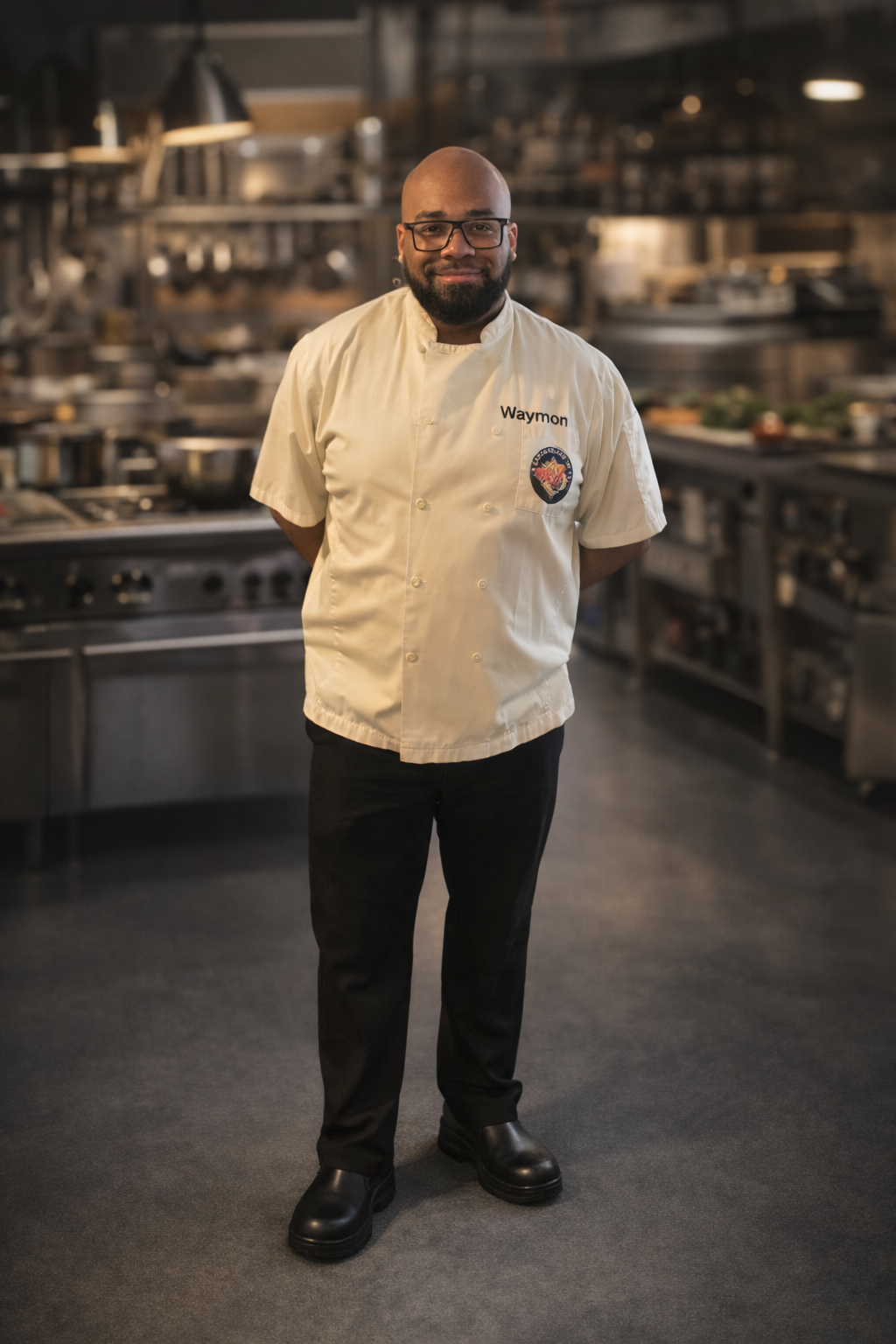 A chef standing in a professional kitchen, wearing a white chef uniform with the name "Waymon" and a patch, smiling with his hands behind his back.