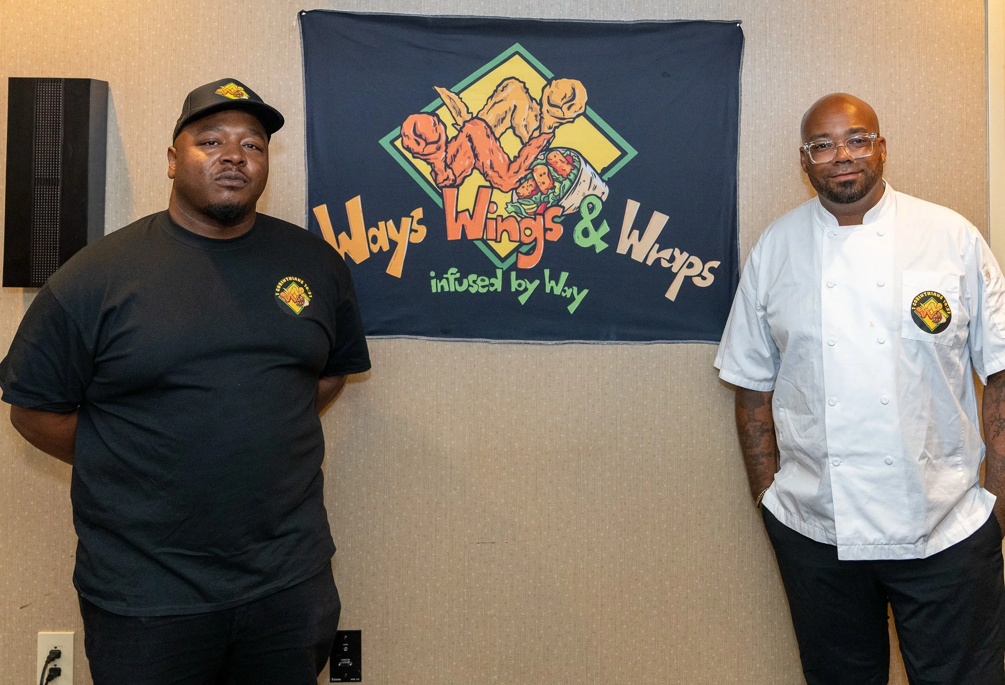 Two men standing on either side of a colorful banner that reads "Wings, Wings & Wraps, infused by Way." The man on the left wears a black t-shirt and baseball cap, and the man on the right wears a white chef's coat with an embroidered logo. The banner features an illustration of wings and wraps in a diamond-shaped design with vibrant colors.