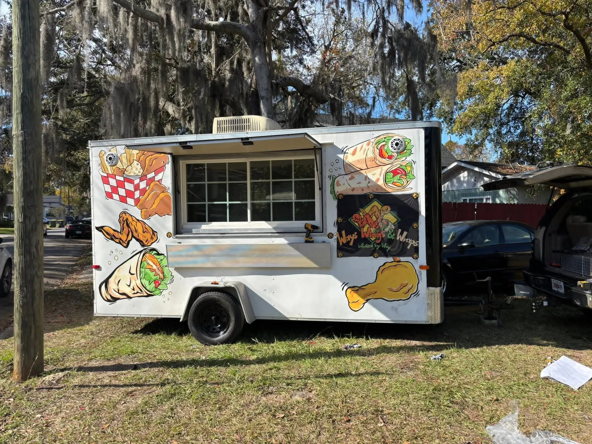 Food truck with illustrations of fried chicken, wraps, and fries on the sides, parked on grass in a neighborhood with trees and houses.