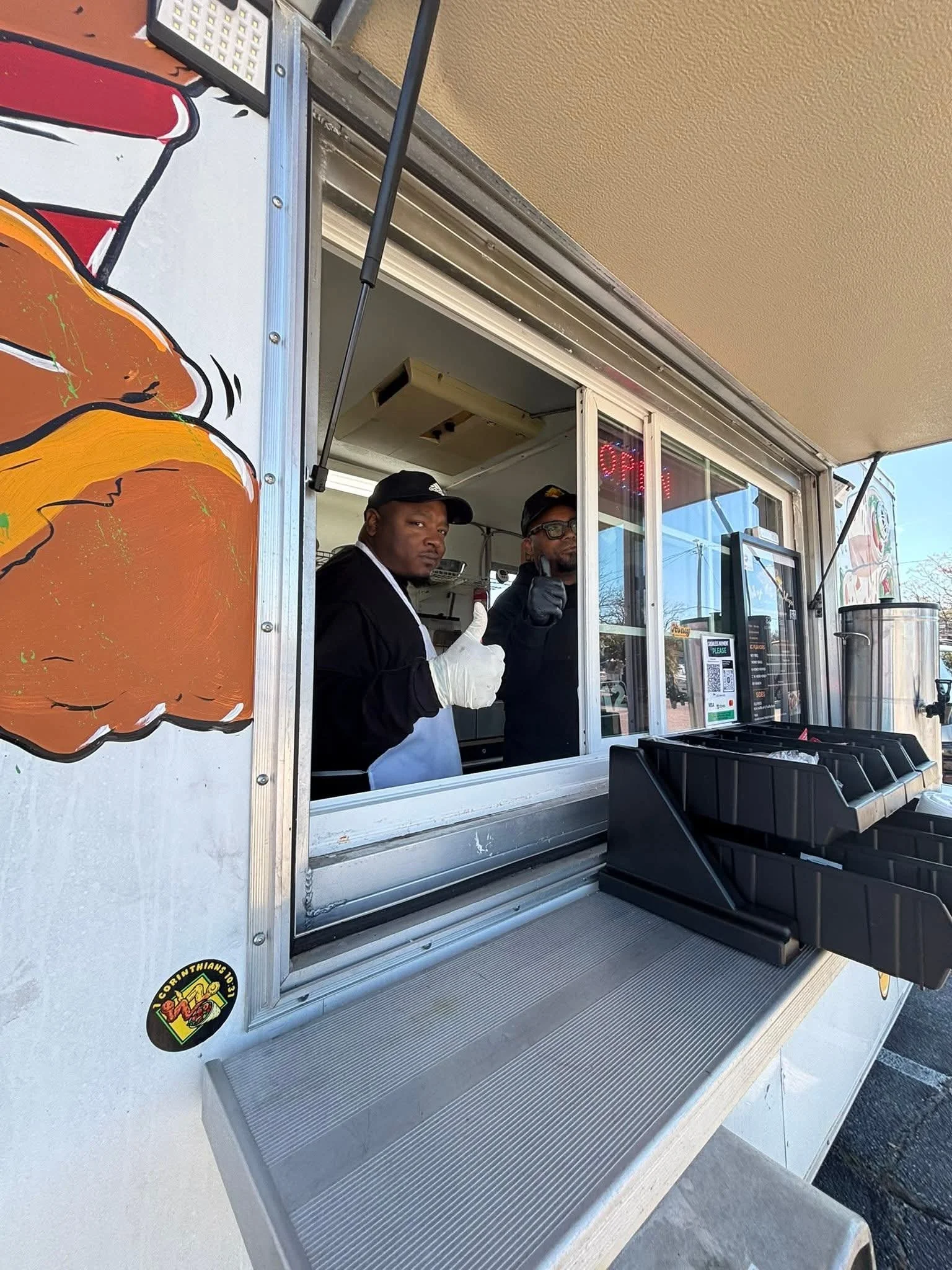Two workers inside a food truck, one showing a thumbs-up and the other giving a wait signal, with an open window displaying parts of the truck's exterior and a menu.