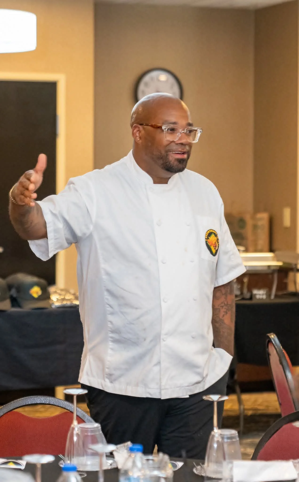 A man in a white chef's coat standing and speaking in a room with round tables and place settings, glasses, and napkins, with a clock and a doorway in the background.