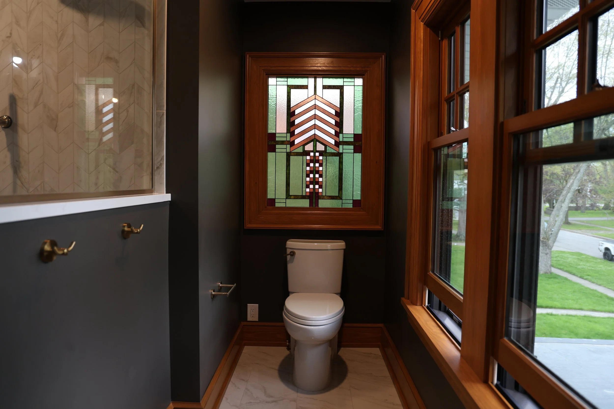 Small bathroom renovation in Amherst, NY with dark walls, featuring a custom stained glass window behind the toilet, wooden trim and large windows to the right showing green outdoor scenery.