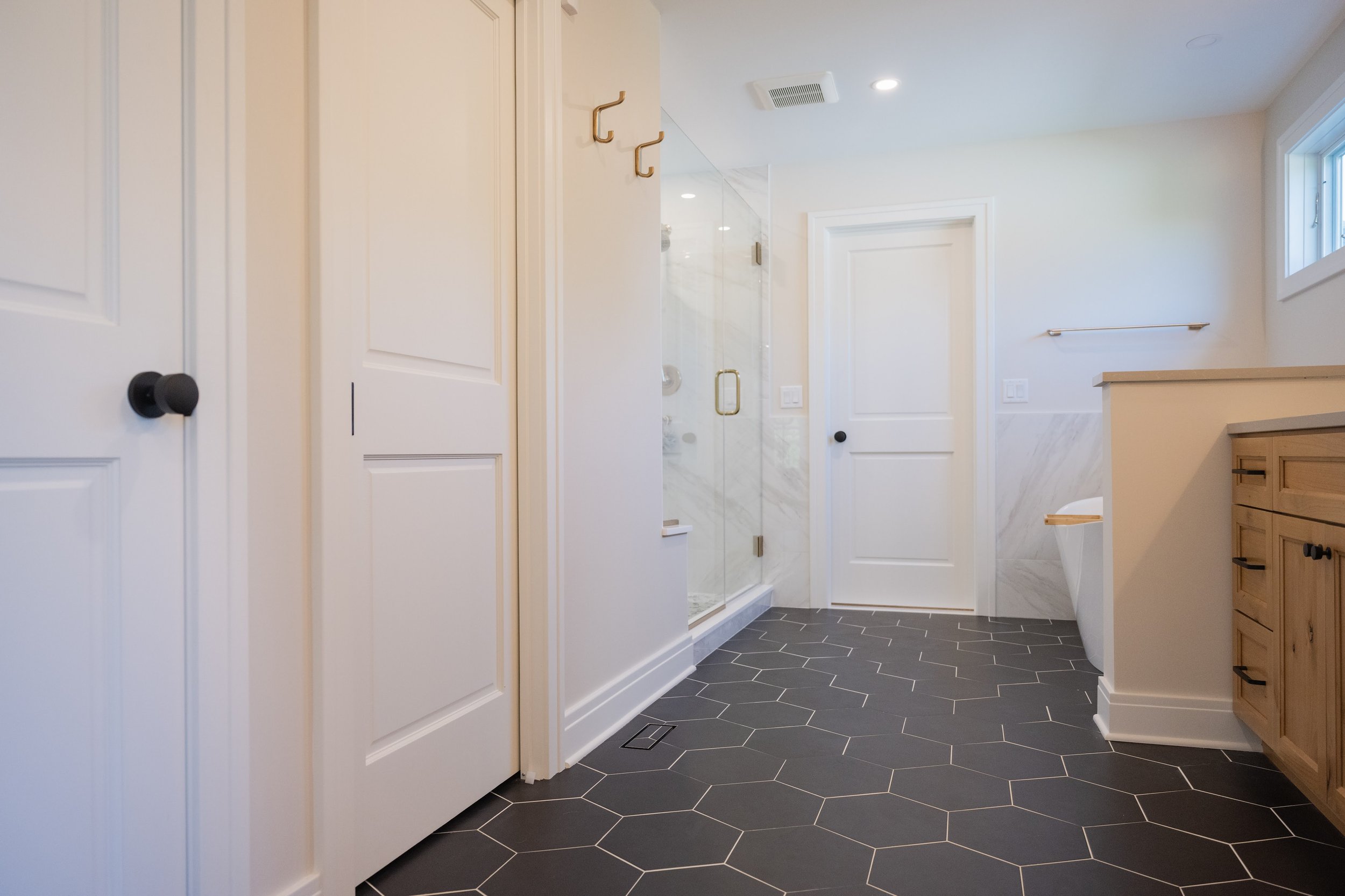 Modern bathroom with black hexagon tile flooring, white walls, a glass shower door, and a wooden vanity.
