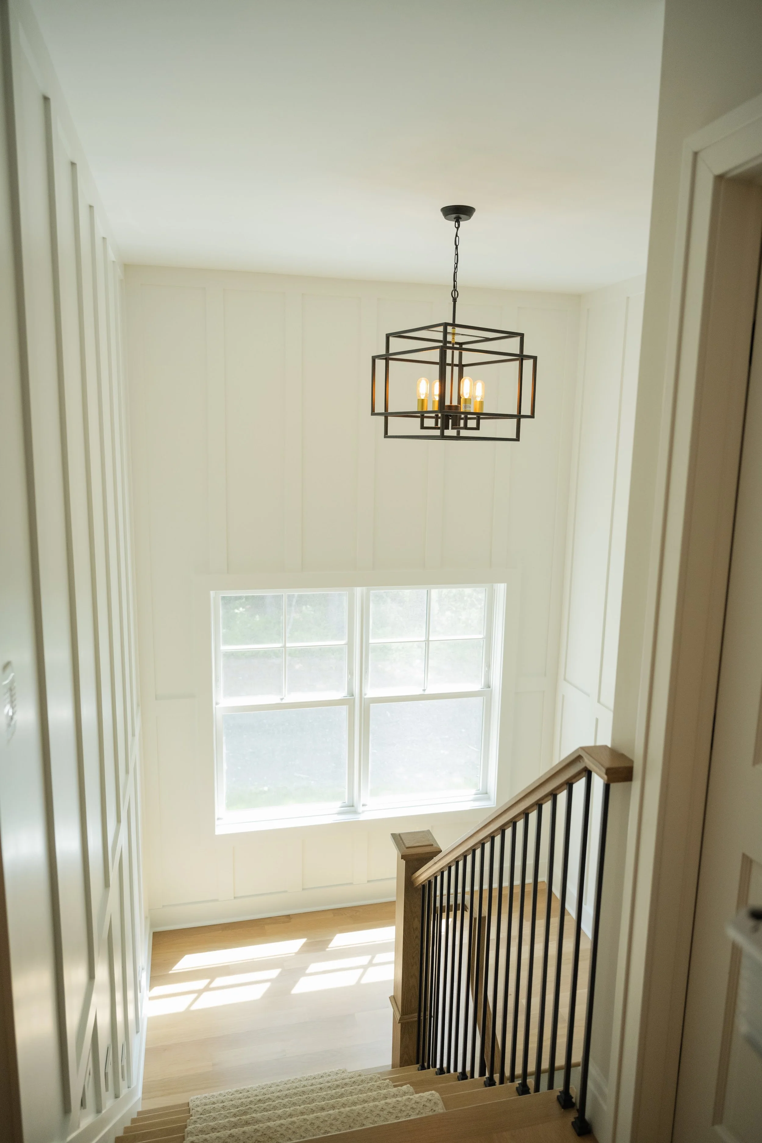 A stairway with a beige carpet leading down, a wooden handrail on the right, and a large window providing natural light in a bright interior space.
