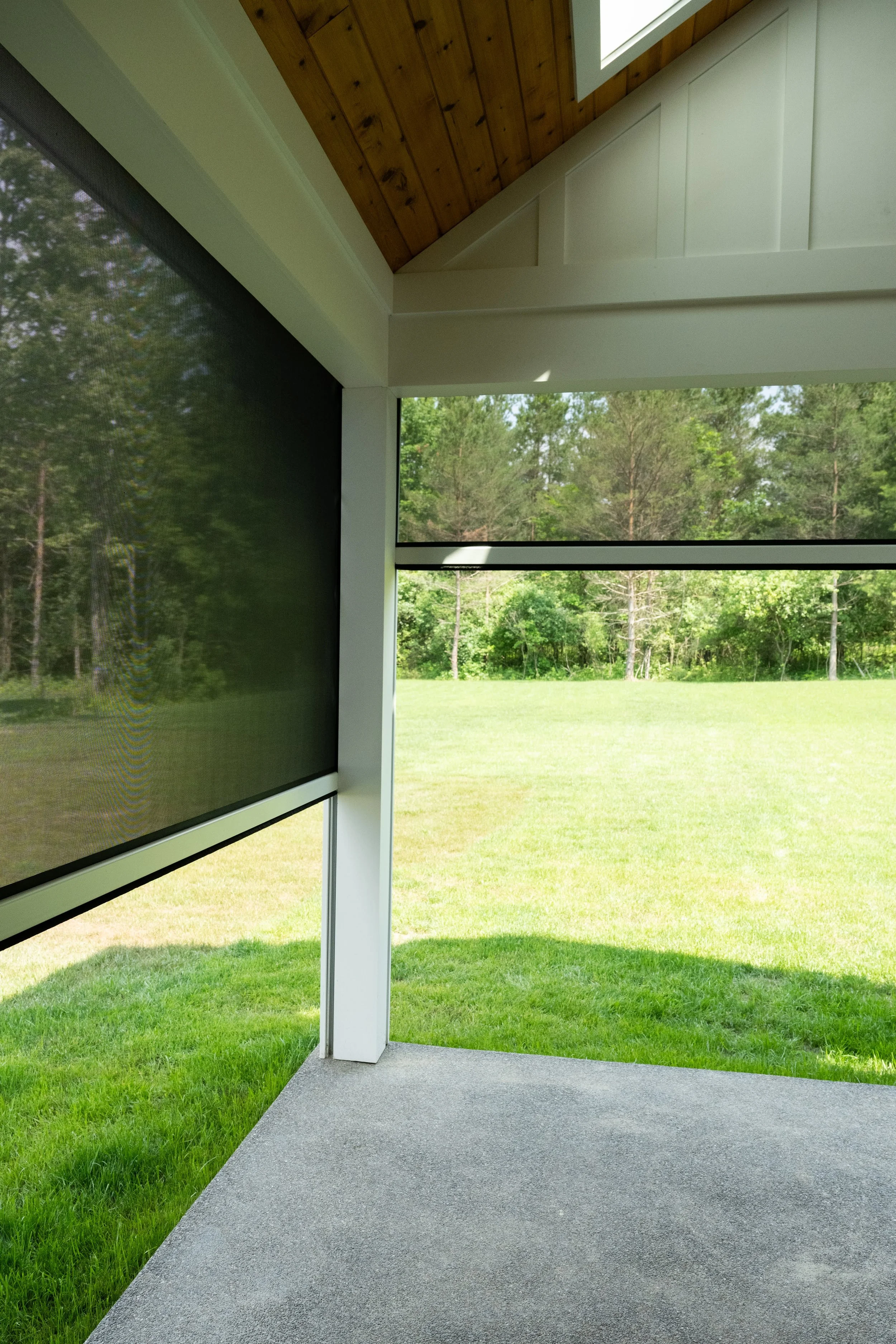 View of a screened porch with a gray carpeted floor and a white supporting column, overlooking a green lawn with trees in the background. The porch has a wooden ceiling and a large window.