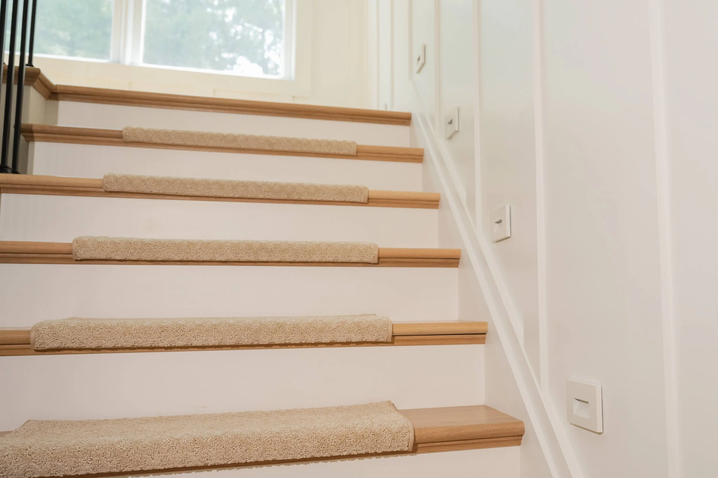 A staircase with beige carpeted steps, wooden trim, and white walls. There is a large window at the top letting in natural light and showing greenery outside. Two electrical outlets or switches are visible on the wall to the right.
