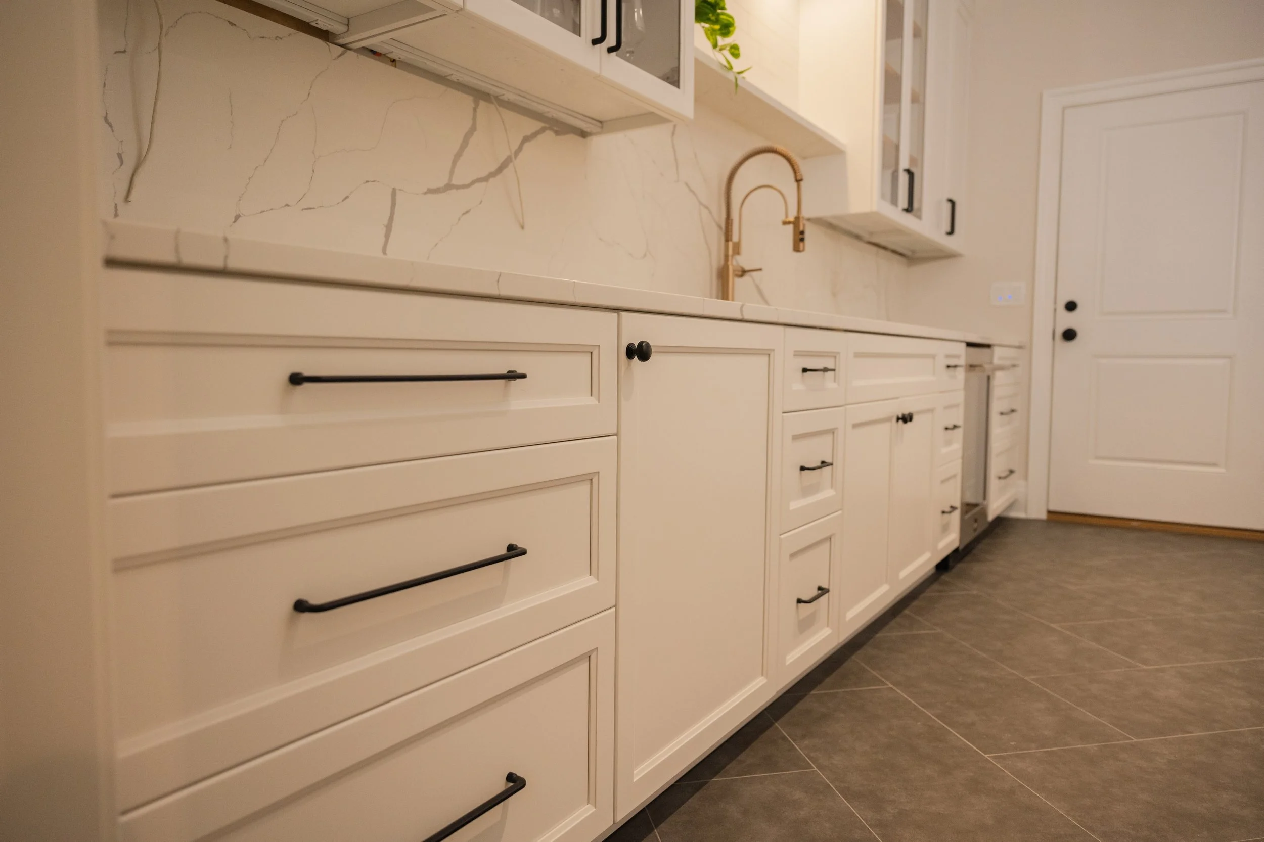 A kitchen with white cabinets, black handles, a gold faucet, and a marble backsplash. Some green plants are visible on top of the cabinets, and there is a dark-colored floor.