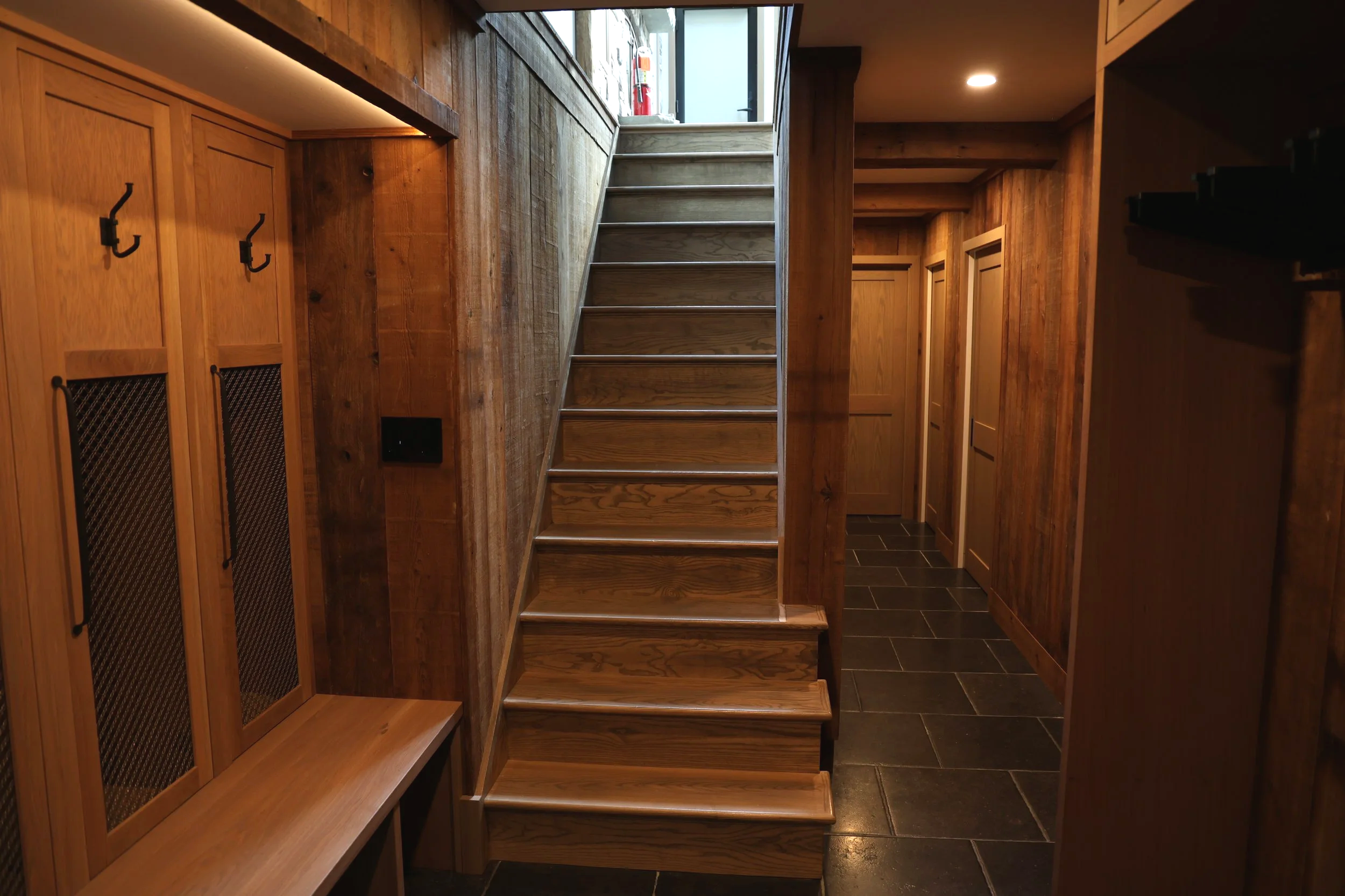 Interior view of a wooden staircase with a narrow hallway, wood-paneled walls, and cabinets with black hooks on the left, leading up to a door with a window. The space is dimly lit with recessed lighting.