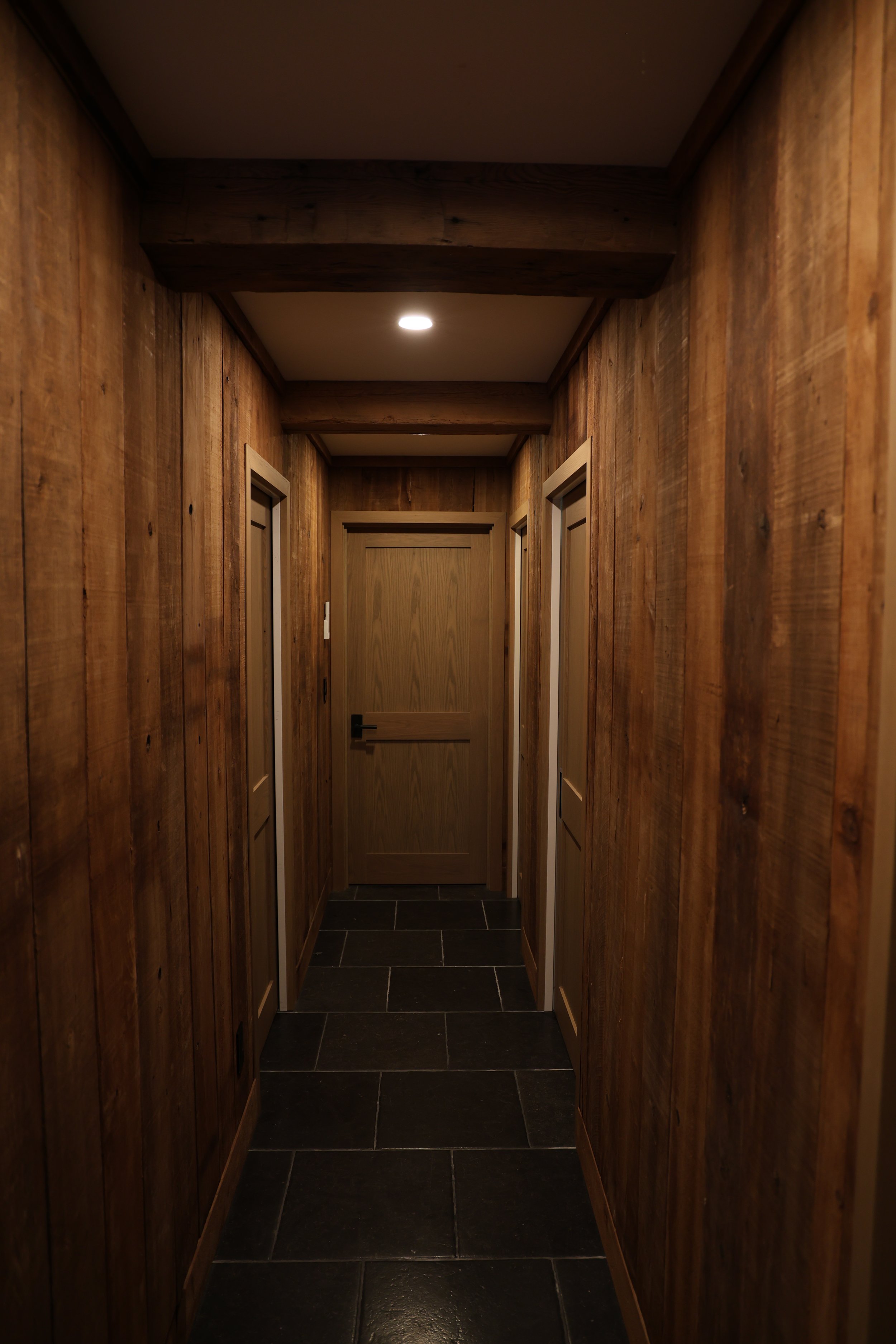Narrow hallway with wooden paneled walls, dark tile flooring, a white ceiling with recessed lighting, and a closed wooden door at the end.
