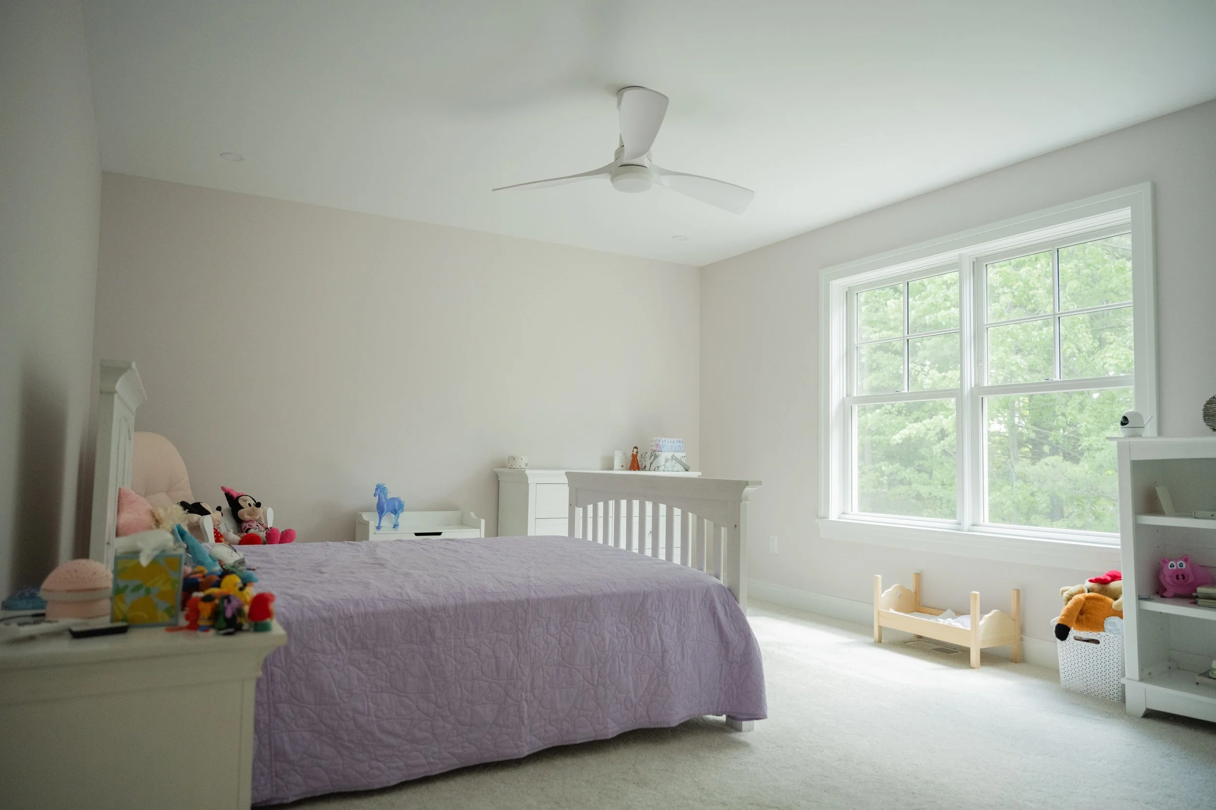 A bright and airy children's bedroom with a white bed and various stuffed animals, including Minnie Mouse and a blue horse, on a white dresser. Large windows with white trim let in natural light, and there is a white ceiling fan. The room has light-c