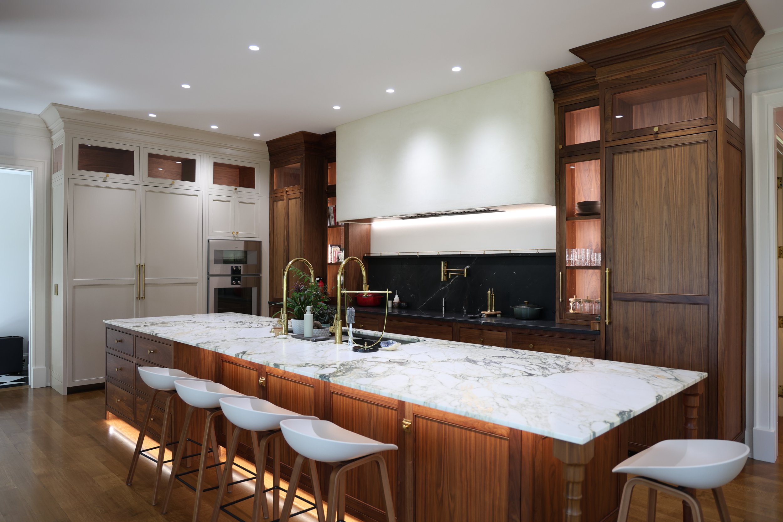 Modern kitchen with white cabinetry, a large marble island with wooden base, brass fixtures, and wooden cabinets with glass accents, under recessed lighting.
