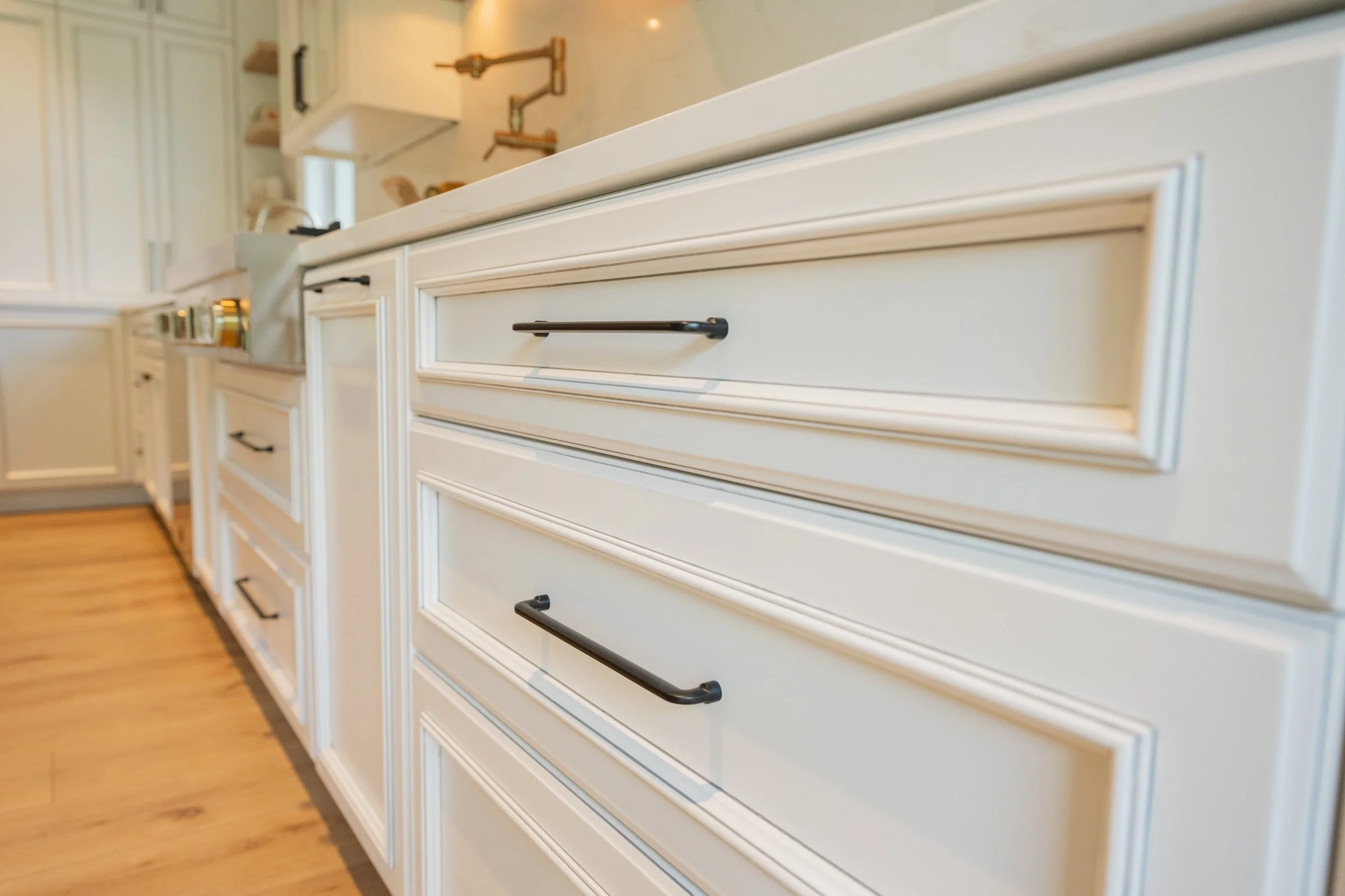 Close-up of white kitchen cabinets with black handles in a modern kitchen with wood flooring.