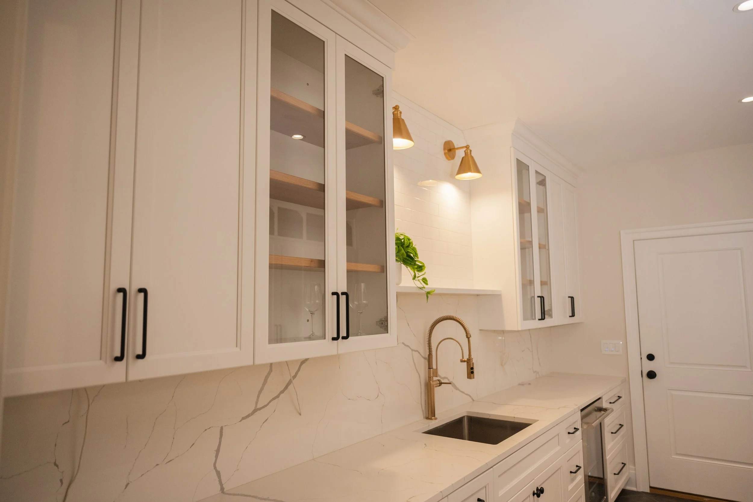 White kitchen cabinets with black handles, a marble countertop with gold veining, a gold kitchen faucet, and gold wall sconces with downward-facing lights.