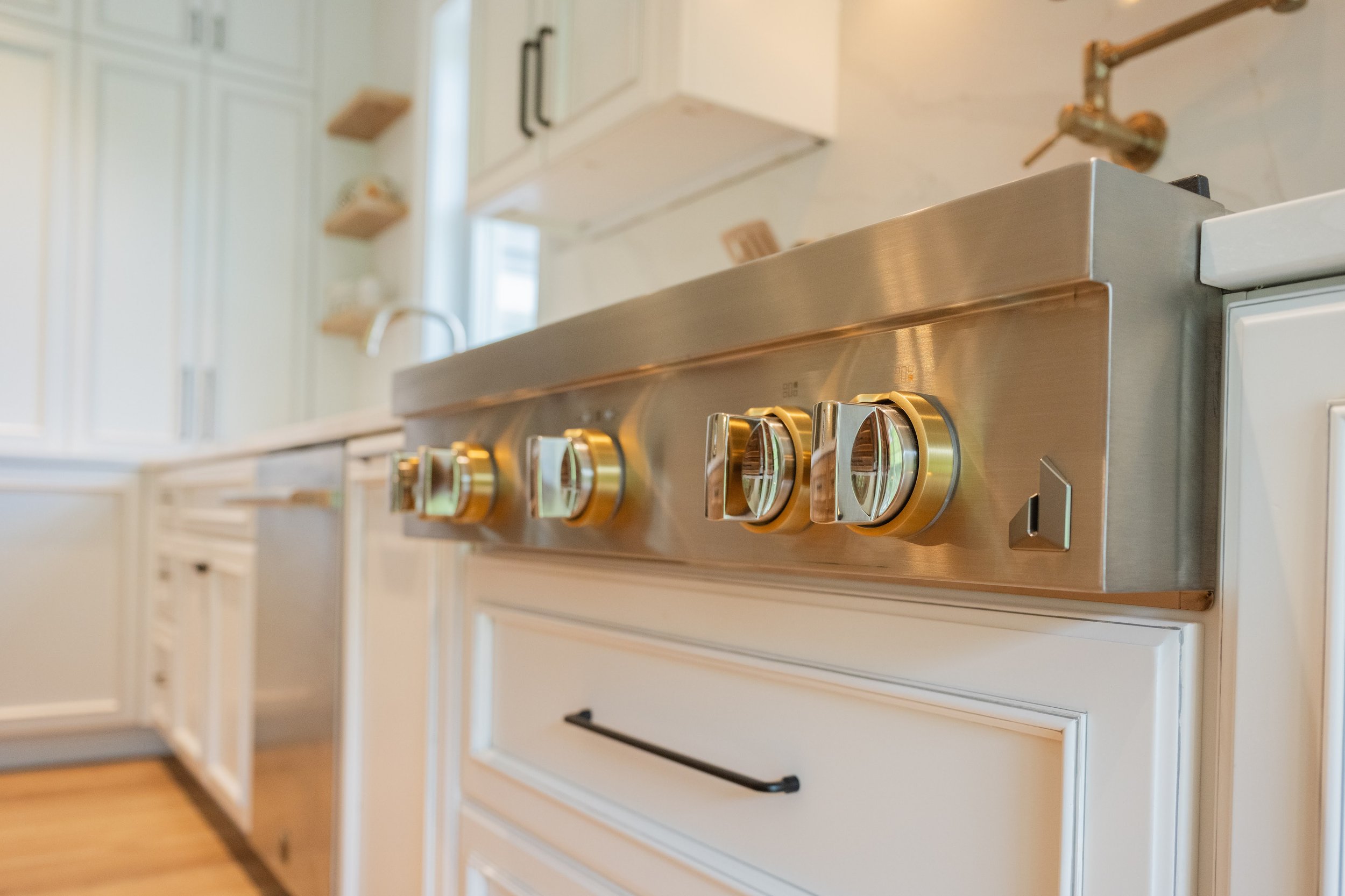 A modern stainless steel gas stove with four control knobs, installed in a white kitchen with cabinets and a window in the background.