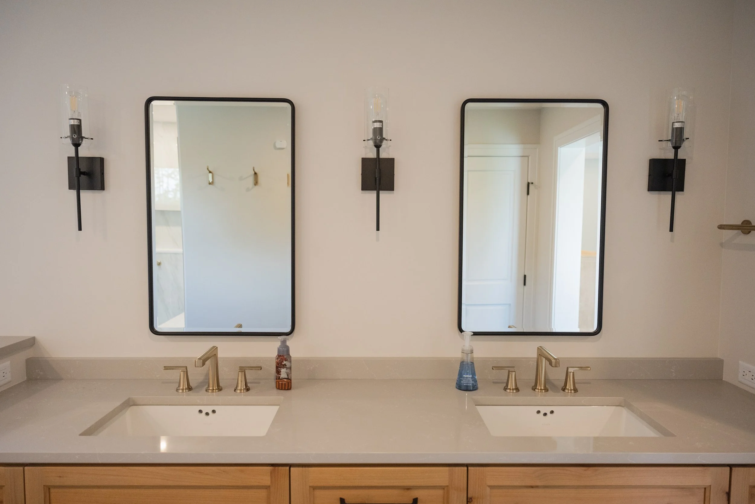 Double bathroom vanity with two rectangular mirrors, black wall-mounted light fixtures, and a beige countertop with a sink, soap dispenser, and a bottle of hand sanitizer. Wooden cabinet below the countertop.