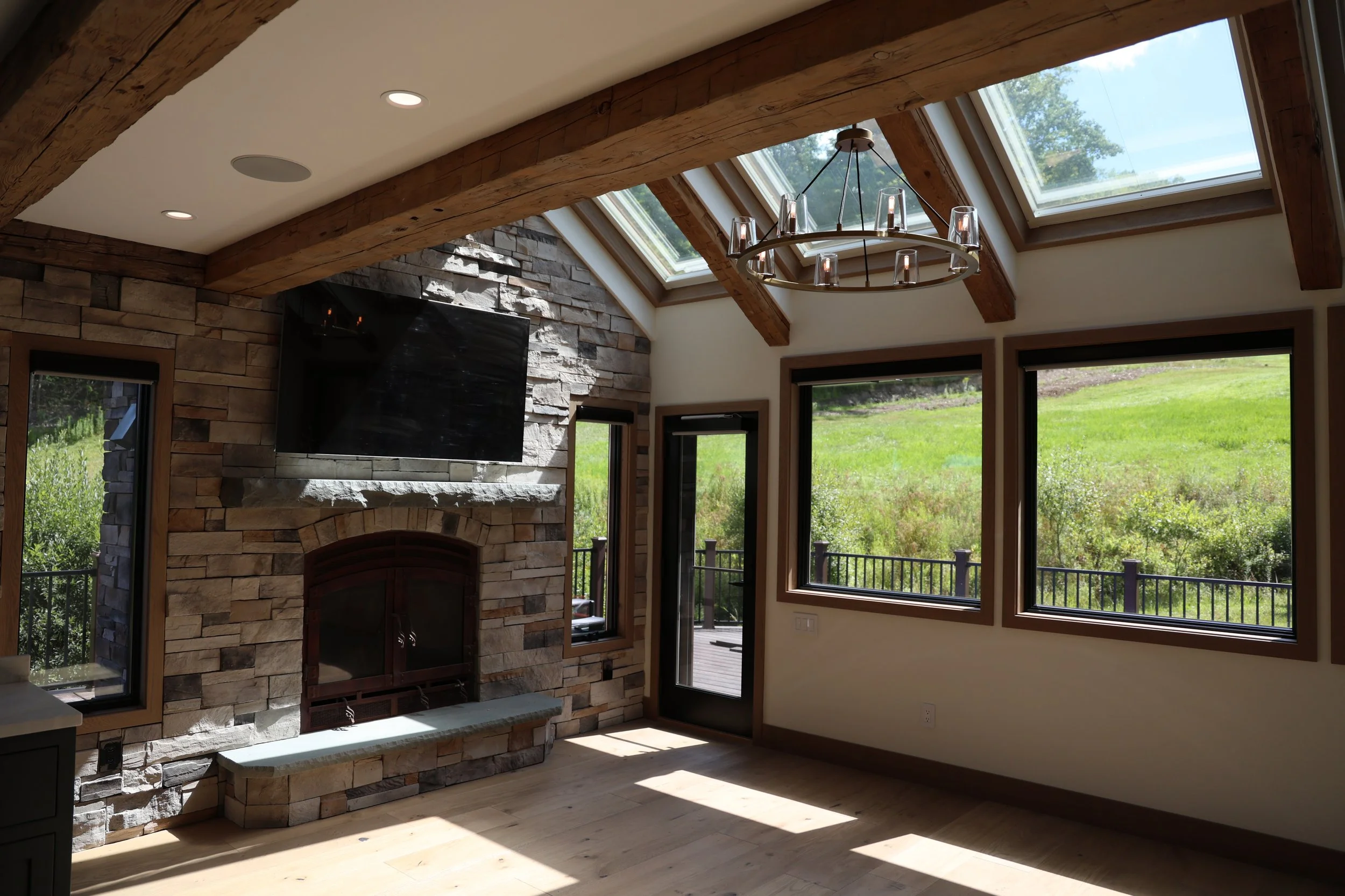 Interior of a living room with a stone fireplace, a mounted flat-screen TV above it, multiple large windows, a sliding glass door leading outside, a chandelier hanging from the ceiling, and a view of a grassy hillside outside.