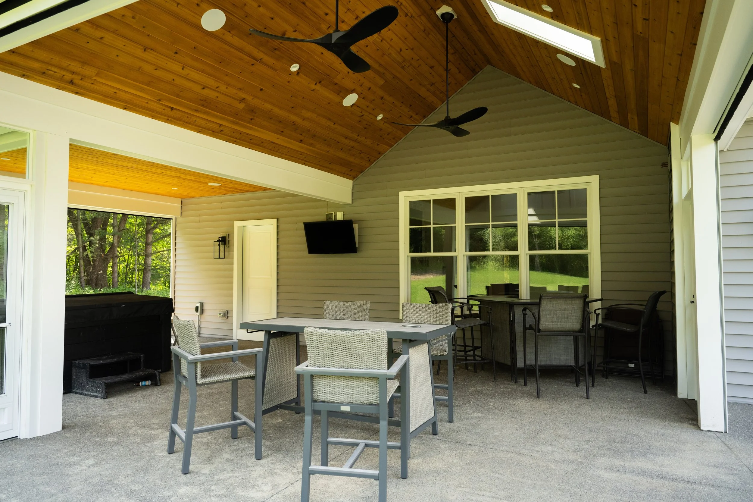 Outdoor covered patio with wicker and metal chairs around a table, ceiling fans, skylights, and large window overlooking greenery.