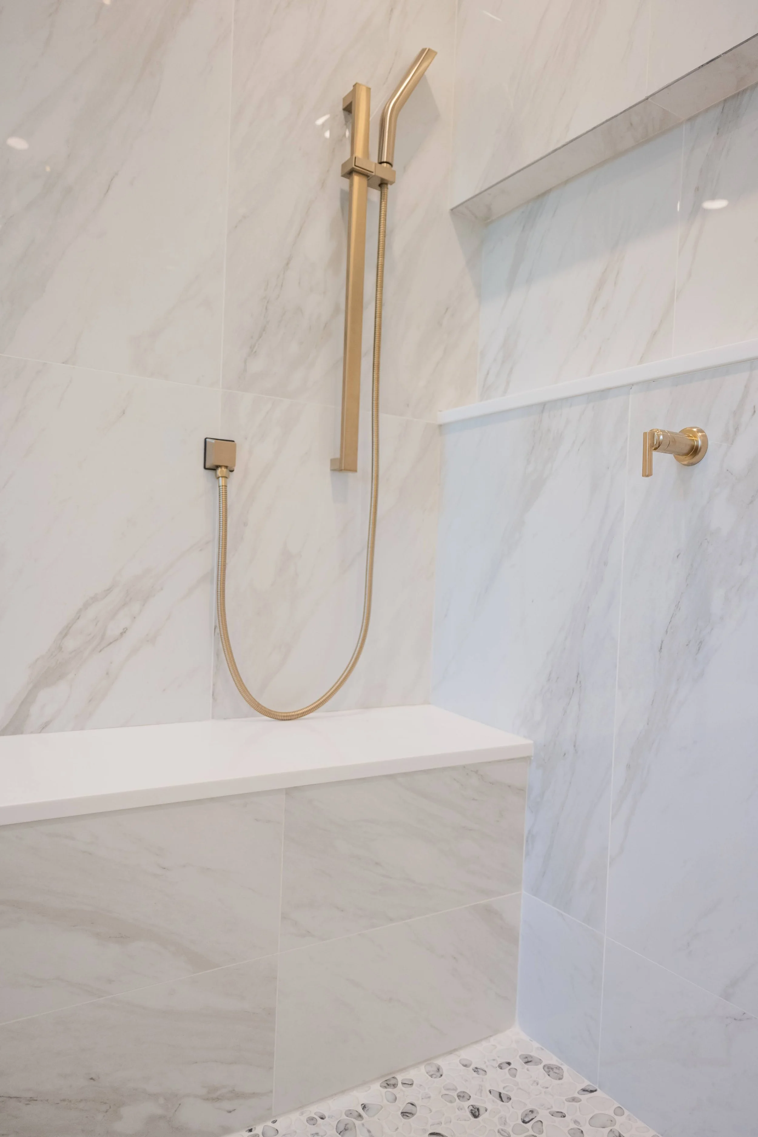 A modern shower with a gold fixtures, a handheld shower head, a built-in shelf, a small towel bar, and a pebble stone floor.
