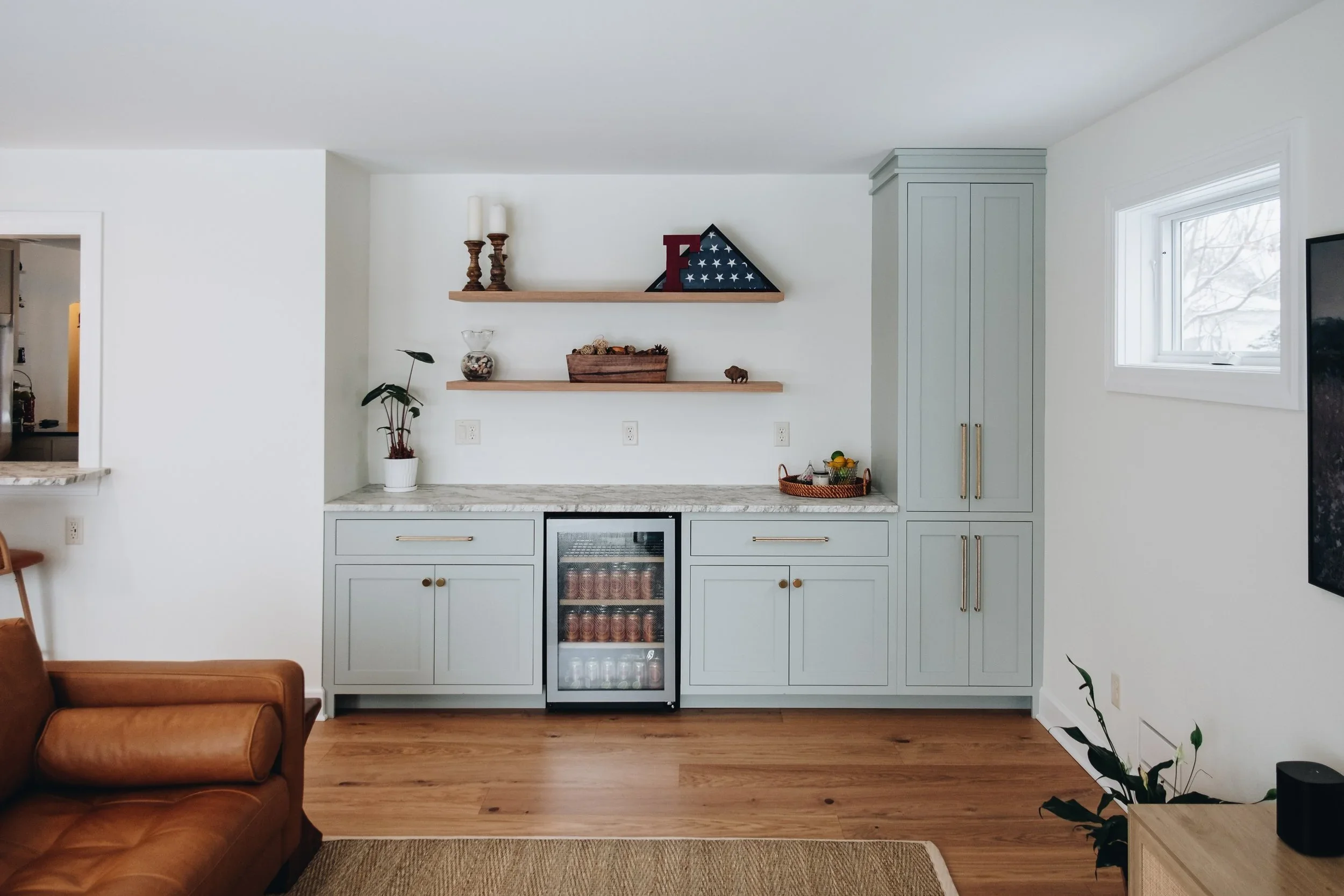A modern kitchen with light blue cabinets, two wooden floating shelves holding candles and decorative items, a small wine cooler, a potted plant, and a window with a view of trees. Part of a leather sofa and a beige rug are visible in the foreground.
