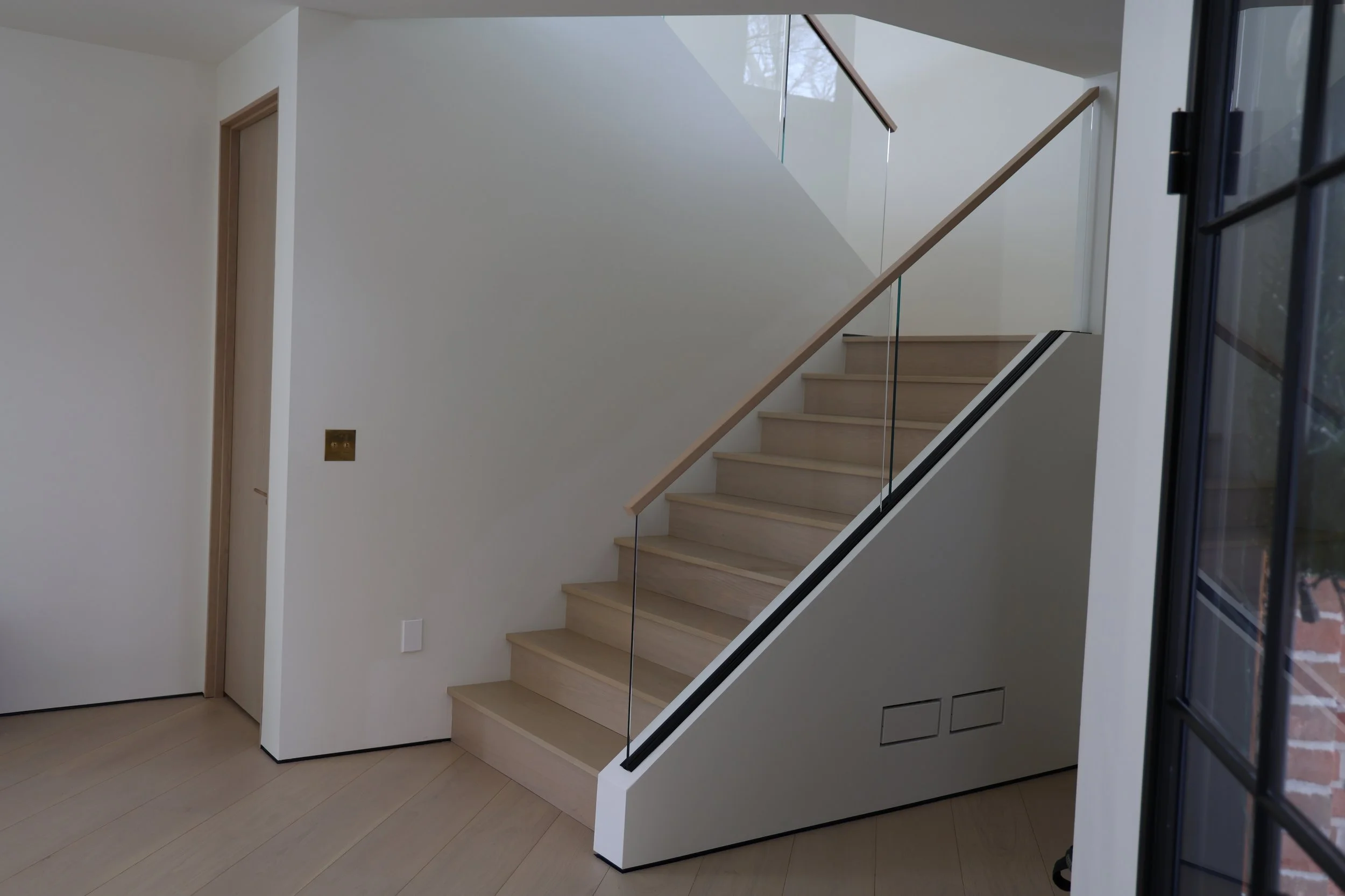 Interior view of a staircase with light wood steps, a glass and wood railing, and a white wall with a door and electrical outlets.