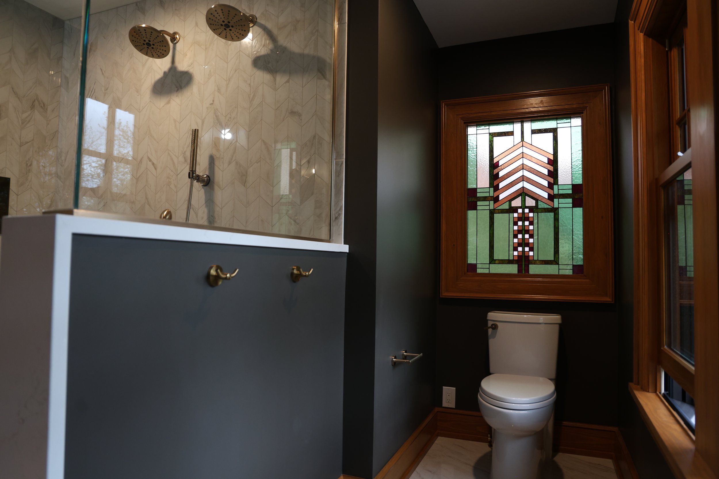 A bathroom with a glass-enclosed shower with two rainfall showerheads, a toilet, a window with stained glass, and dark painted walls with wooden trim.
