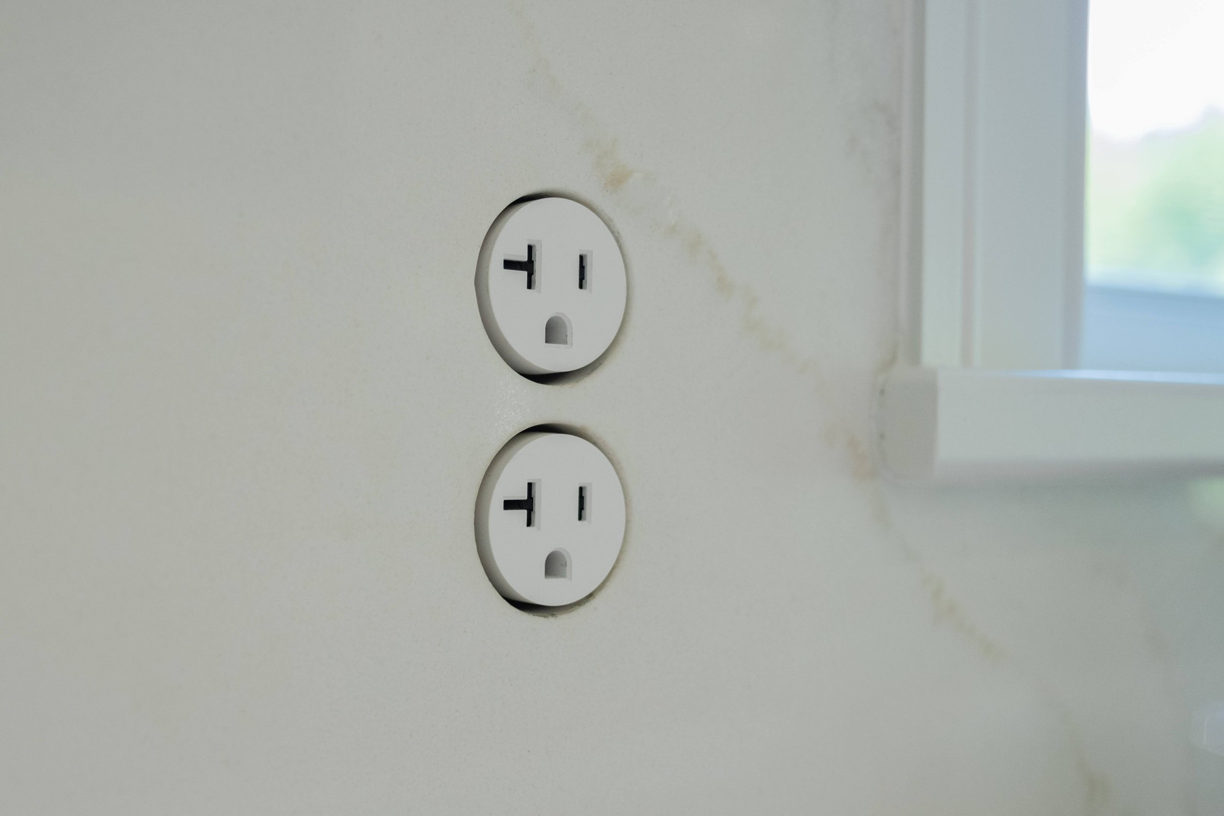 Close-up of electrical outlets on a white wall with visible dirt and stains, next to a window with blurred exterior view.