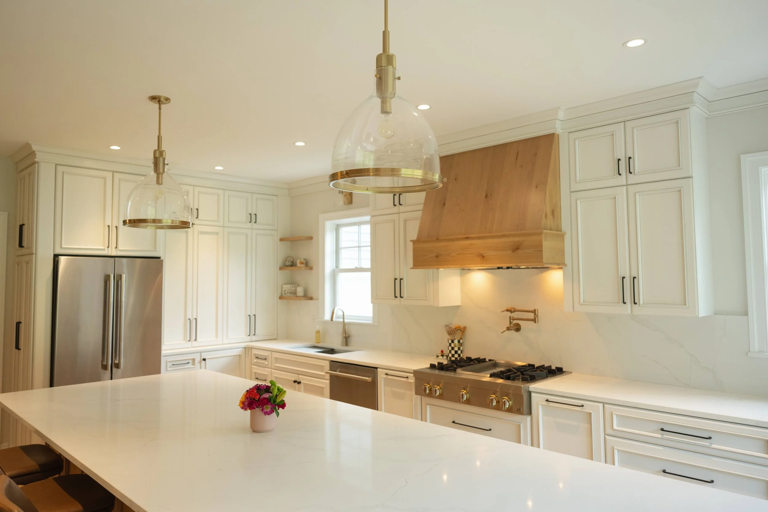 Modern white kitchen with pendant lights, a large kitchen island with a pink flower arrangement, stainless steel refrigerator, and wooden accents on the range hood.