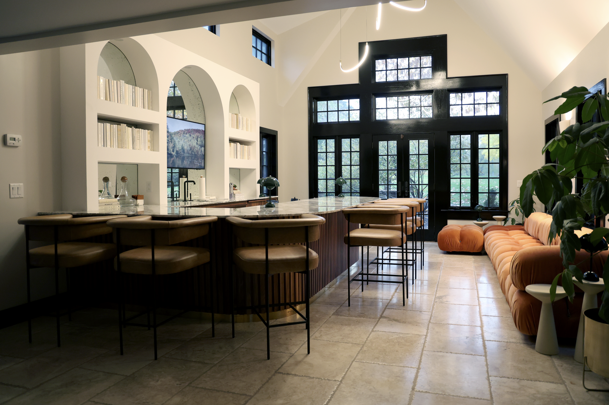 Modern indoor living space with a large black-framed glass door, a high ceiling, a built-in bookshelf with white arch openings, a flat-screen TV, a marble countertop, beige bar stools, orange velvet sofa, and a green potted plant.